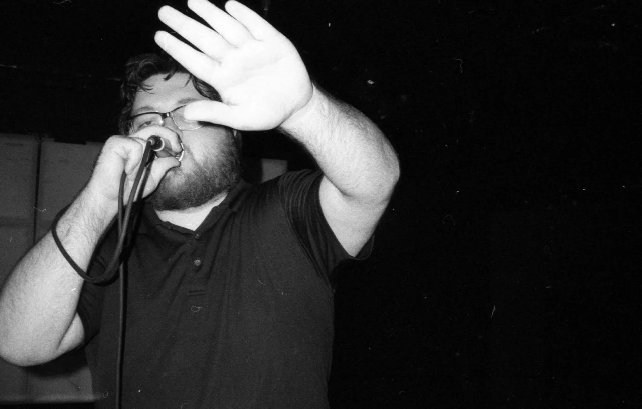 A man with glasses and a beard singing into a microphone with his hand raised, wearing a black shirt.