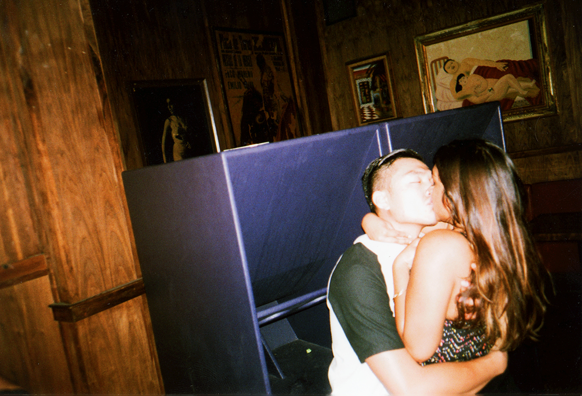 A young couple sharing a kiss in a dimly lit room with wooden walls and framed artwork.