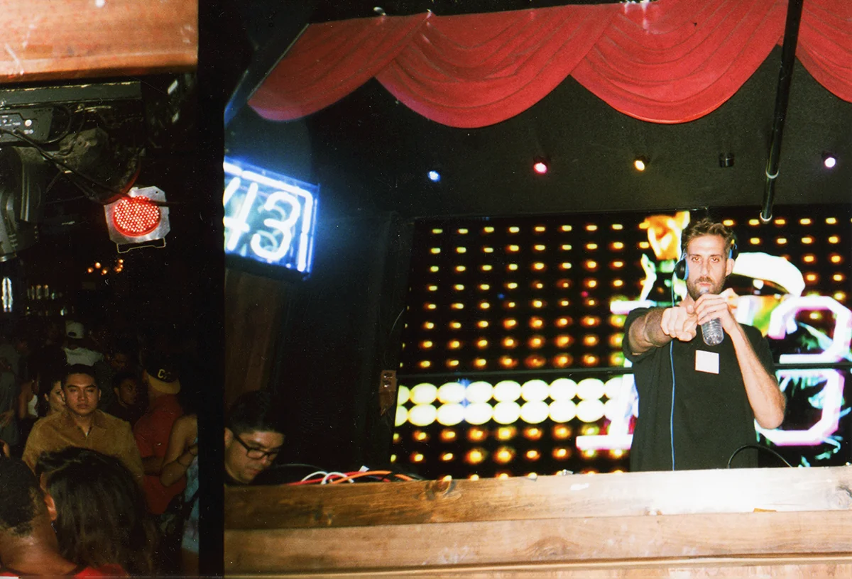 A DJ is playing music at a nightclub with a crowd watching. The DJ is holding a water bottle and wearing headphones. The background has bright LED lights and a red curtain above.