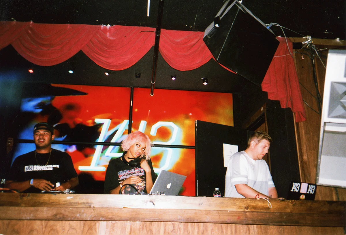 Three DJs with equipment, standing behind a wooden DJ booth in a club or music venue, with a red and black backdrop and large screen displaying '120' in the background. 143