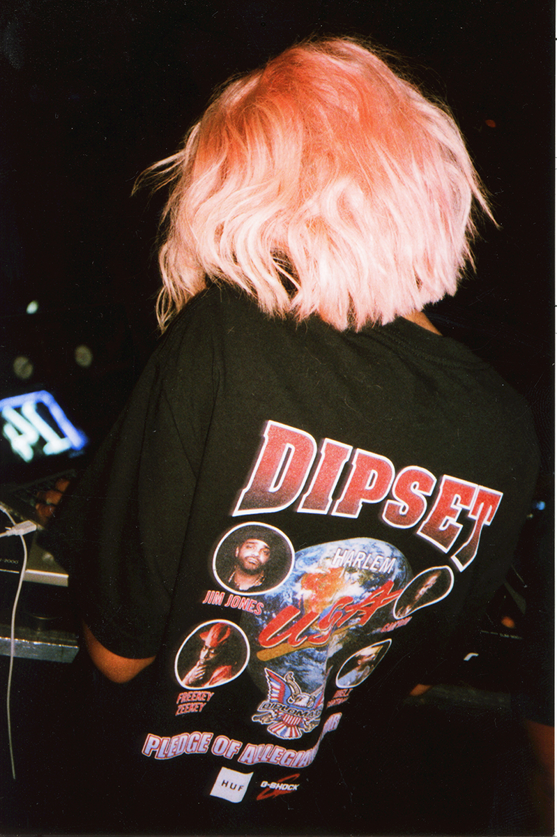 Back of a person with pink hair wearing a black T-shirt featuring a vintage-style graphic design with the word DIPSET, images of various artists, and slogans.