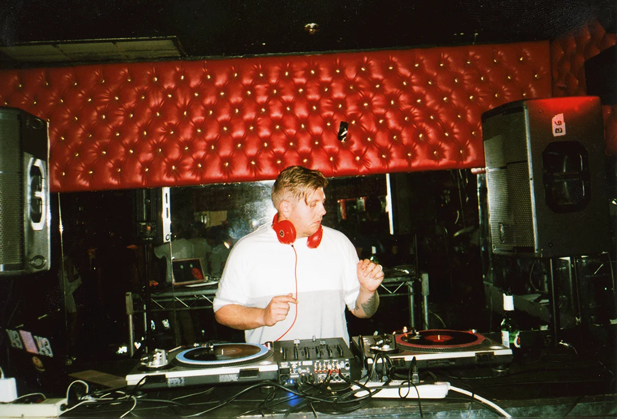 A DJ with blonde hair wearing a white shirt and red headphones is playing music at a club with a red, tufted leather wall in the background. There are DJ equipment and speakers on either side.