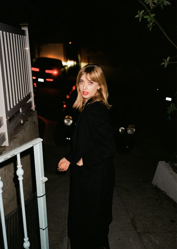 A young woman with shoulder-length blonde hair and red lipstick standing outdoors at night, wearing a black coat and pants, with cars parked in the background.