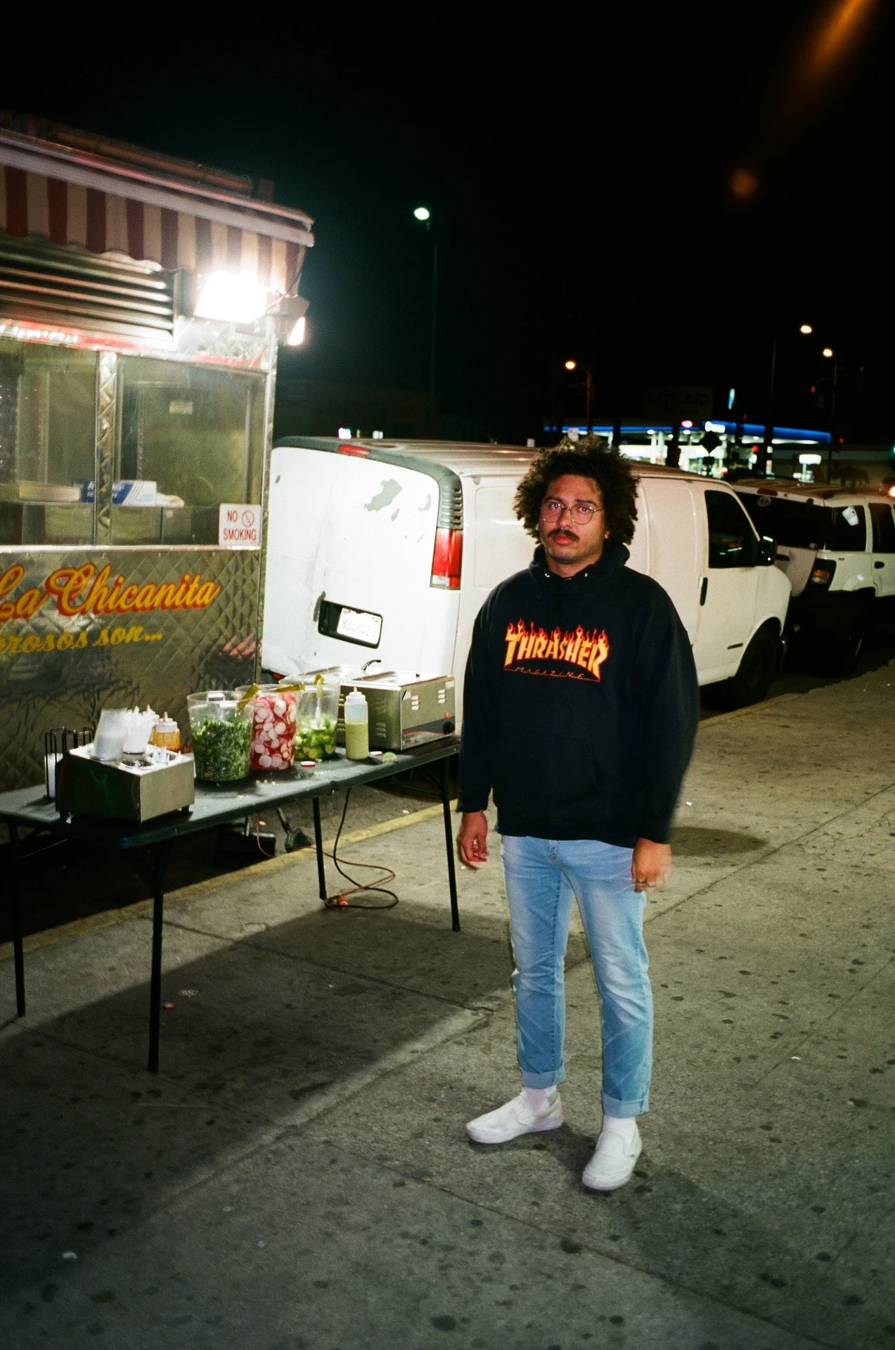 A man standing on a sidewalk at night next to a food stand. The stand has various ingredients and condiments on a table in front of it. The man is wearing a black hoodie with a flaming 'Thrasher' logo, light blue jeans rolled up at the cuffs, and whi