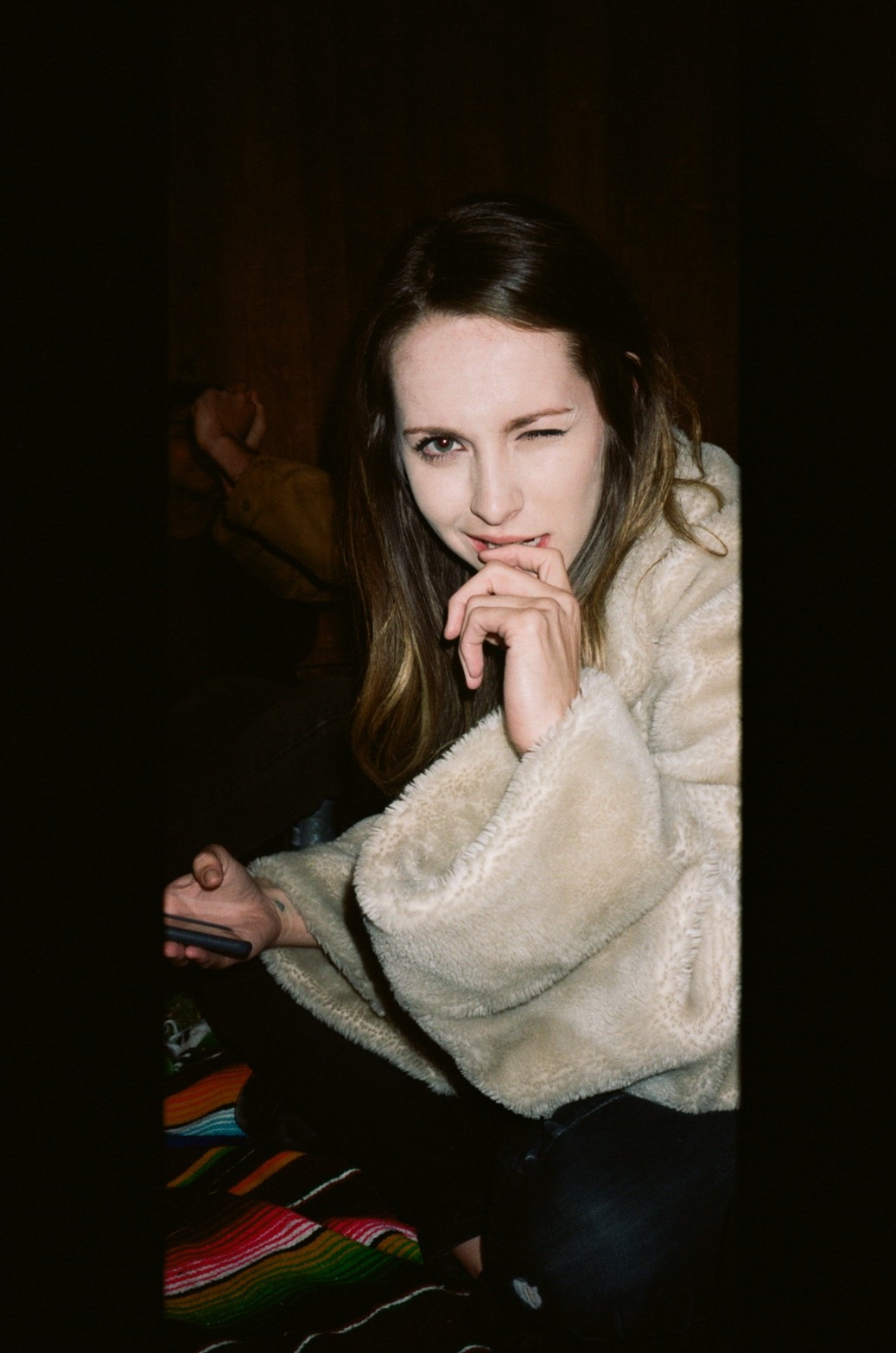 A young woman with long brown hair, fair skin, and wearing a fluffy beige jacket, making a playful expression with one eye winking and her hand near her mouth, sitting at a table with a colorful striped tablecloth, in a dimly lit room.
