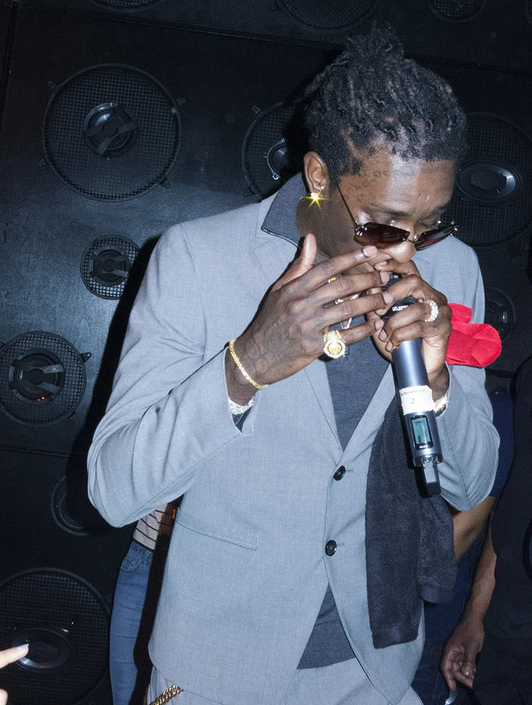 A man wearing a gray suit, sunglasses, and jewelry is holding a microphone and appears to be singing or speaking at an indoor event, with black speakers in the background. music, editorial, asap ferg