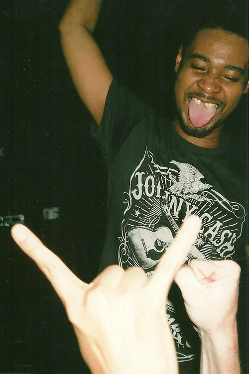 A young man with short hair and a goatee sticking out his tongue, smiling, wearing a black t-shirt with a graphic of a guitar and text. Two hands in the foreground making rock and roll signs with their fingers. Danny Brown, music, editorial 