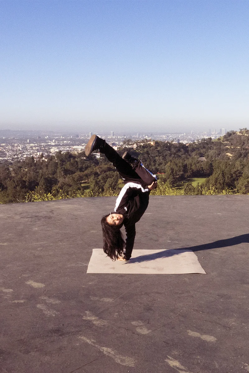 A woman performing a handstand with her legs extended upward on a yoga mat outdoors, with a city skyline in the background and trees on a hillside.