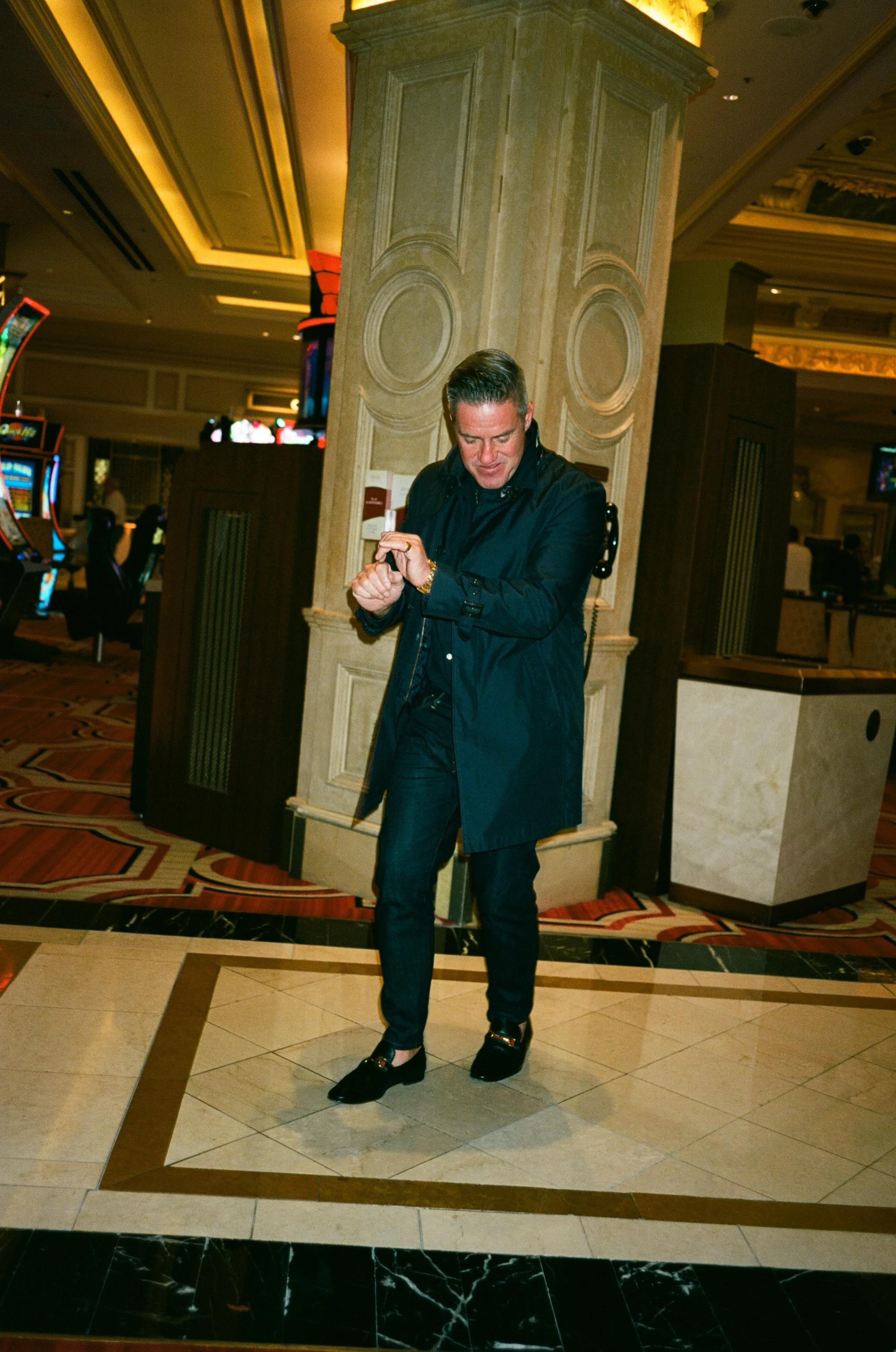 A man in a black jacket and dark jeans standing on a tiled floor in a casino, looking at his watch.