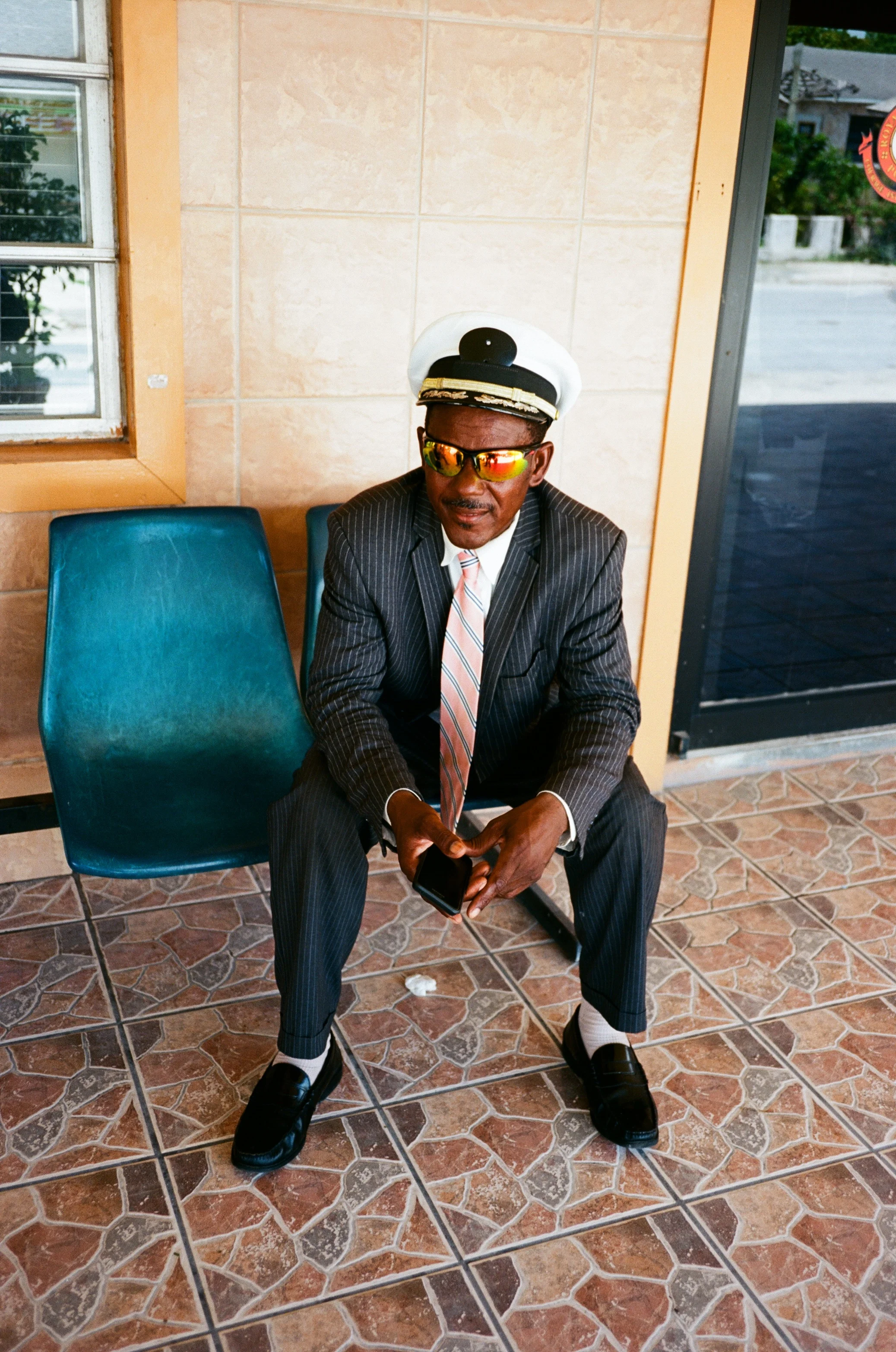 A man dressed in a pinstripe suit, pink tie, and white shirt, sitting on a green plastic chair. He is wearing a white captain's hat, reflective sunglasses, and black loafers with white socks. He is holding a smartphone in his hands and the setting ap