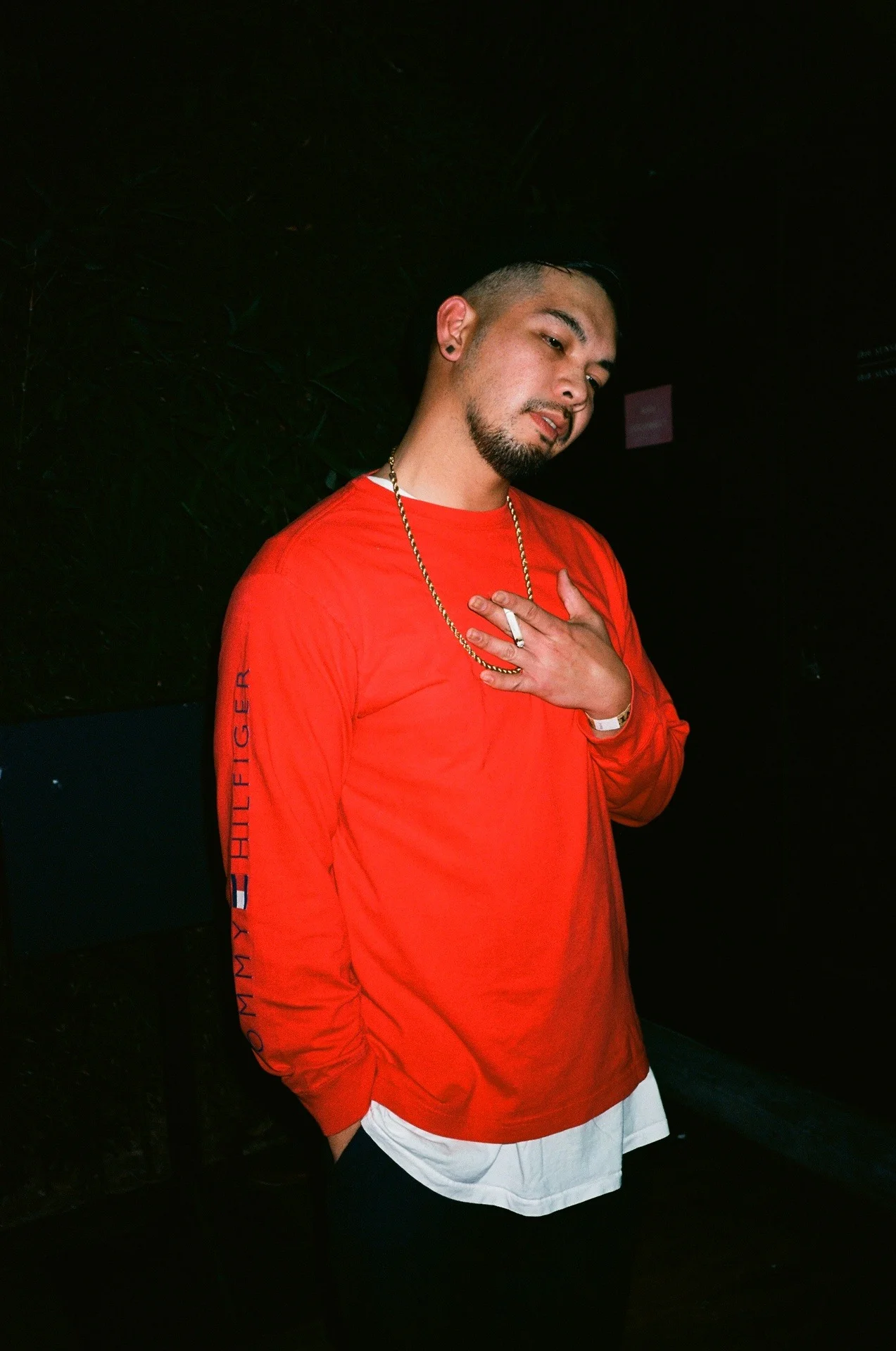 A young man with a goatee and earrings wearing a red long-sleeve shirt, a gold chain, and a white shirt underneath, standing against a dark background.