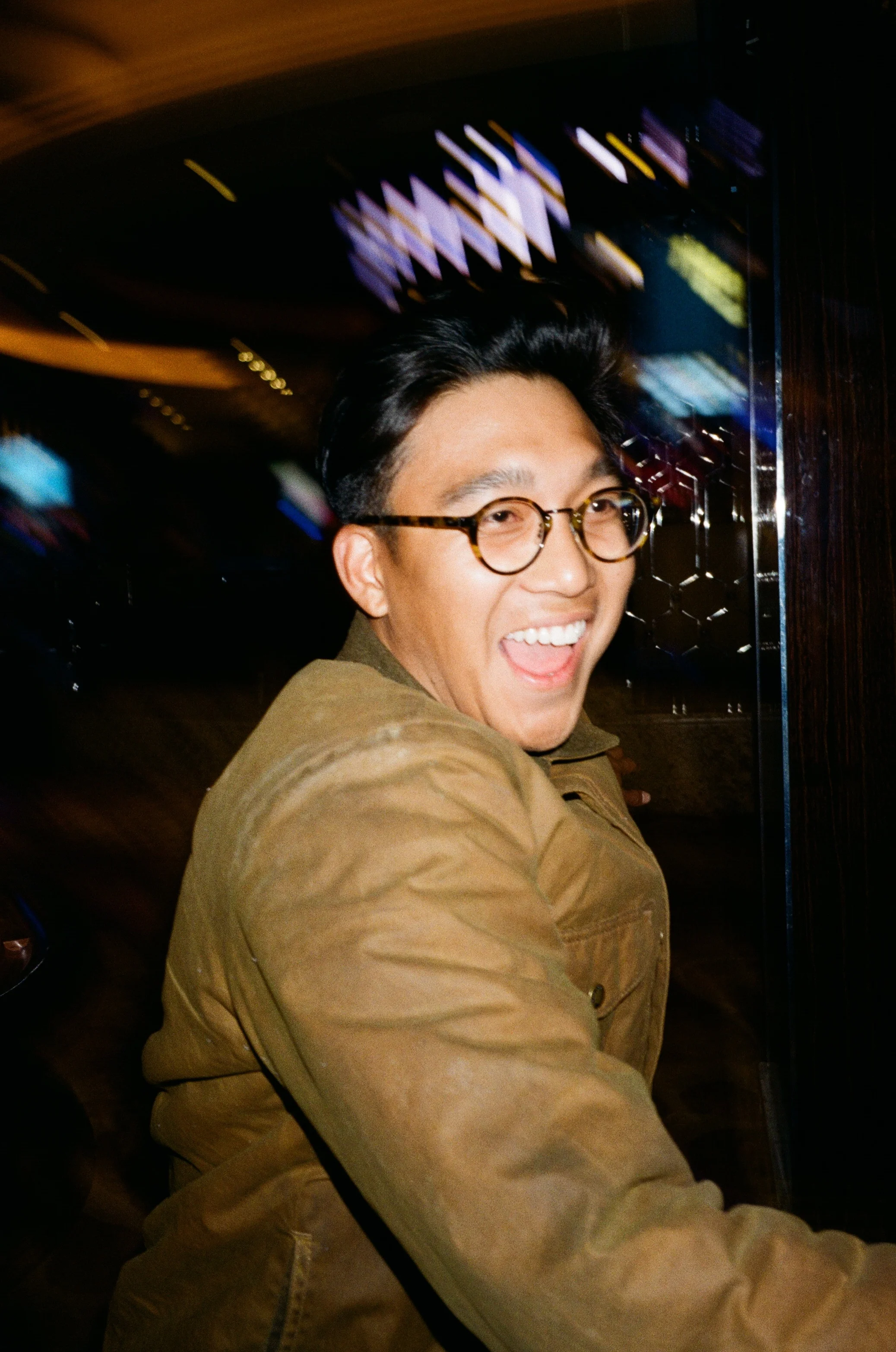 A young man wearing glasses and a brown jacket, smiling and facing the camera in a dimly lit environment with blurred colorful lights in the background.