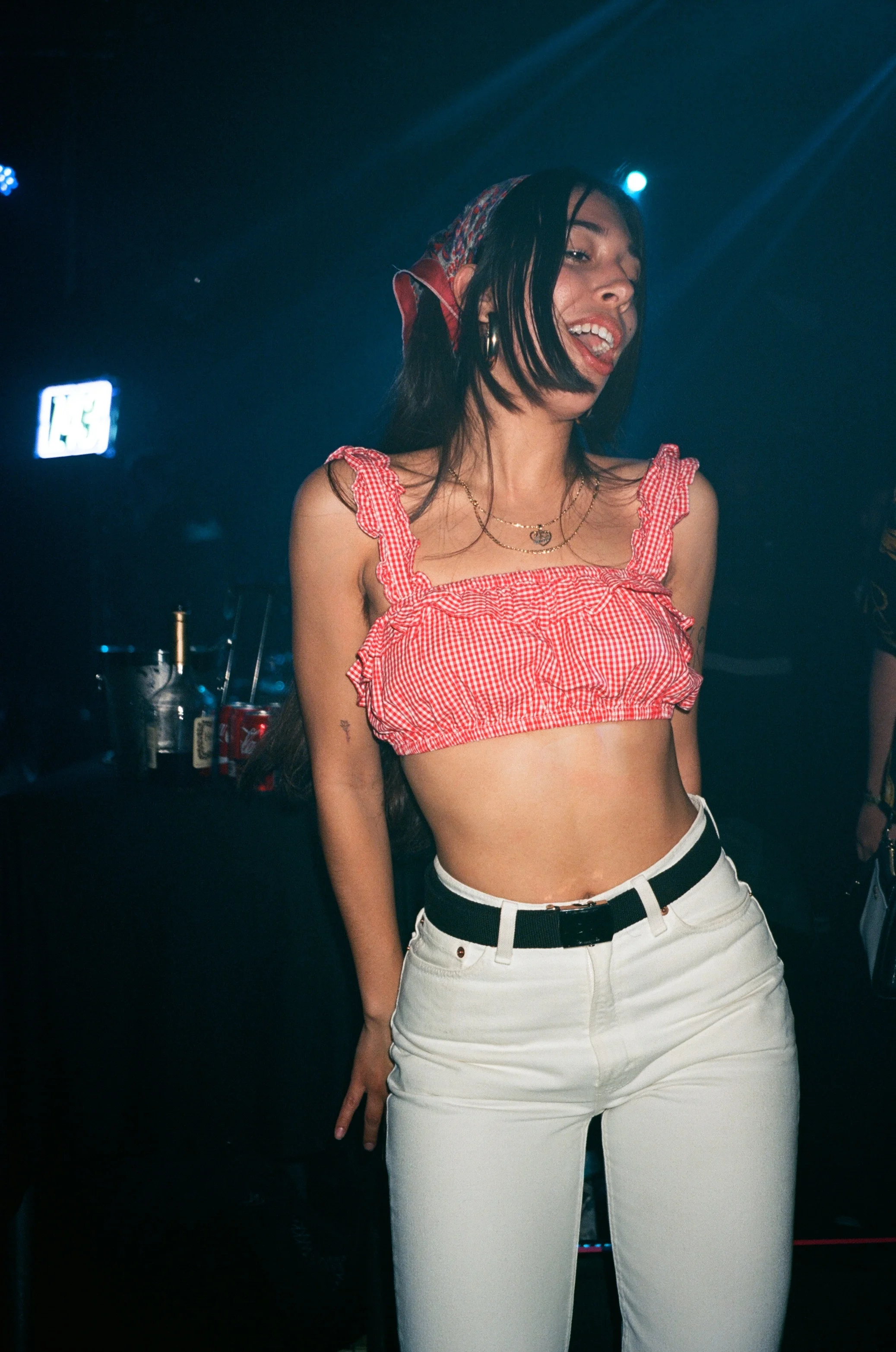 Young woman with long dark hair wearing a red checkered crop top, white pants, and a black belt, dancing in a dark club with blue lighting. music, editorial, nightlife 