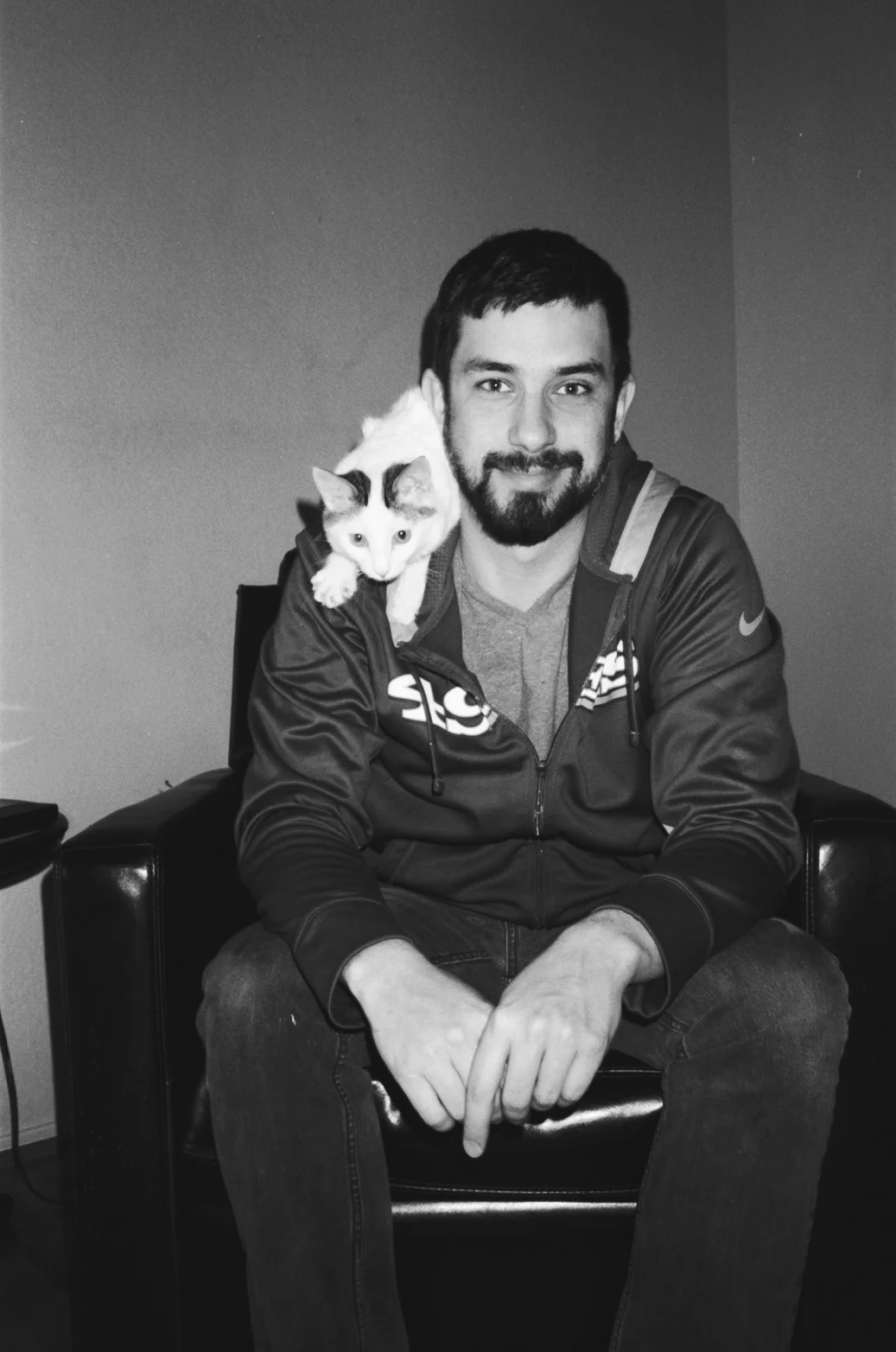 A black and white photo of a man sitting on a couch with a cat perched on his shoulder.