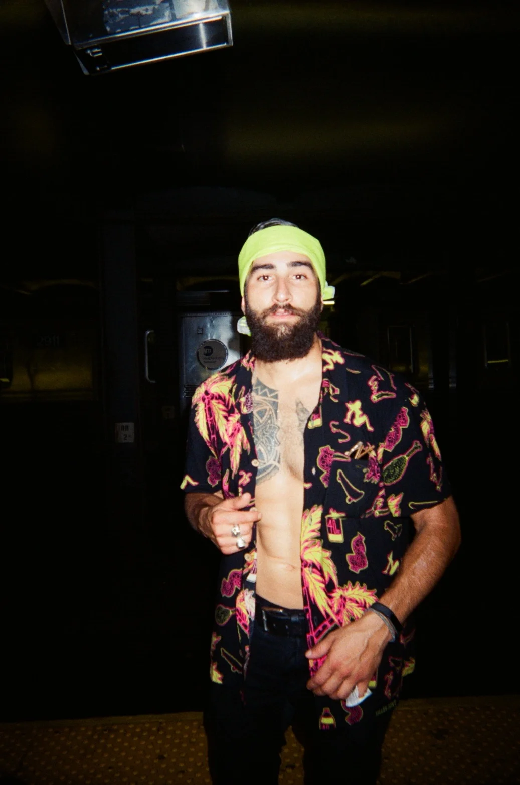 A man with a beard wearing a yellow headband and an open, colorful Hawaiian shirt revealing a tattoo on his chest, standing in a dark indoor space.
