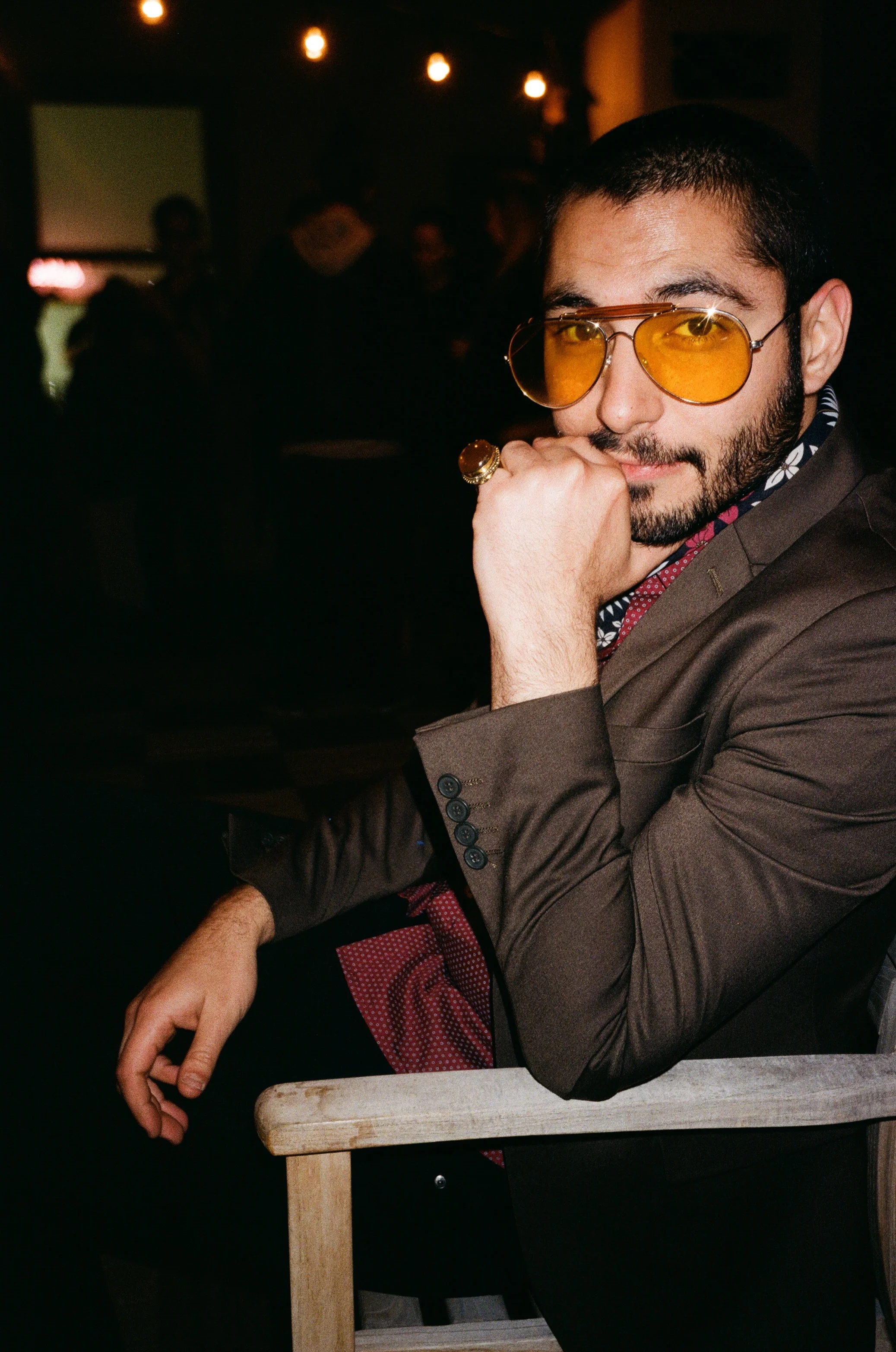 A man with a beard wearing yellow-tinted sunglasses, a brown blazer, and a patterned shirt, sitting at a table in a dimly lit setting.