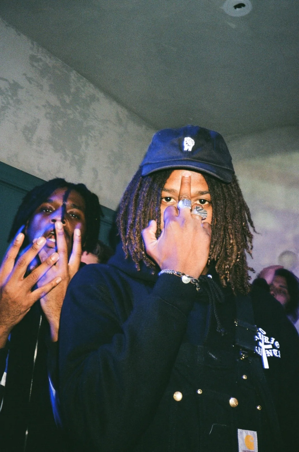 Two young men pose with intense expressions; one is wearing a black cap and jewelry, making a middle finger gesture, and the other is holding up three fingers near his face at the background of a dimly lit room.