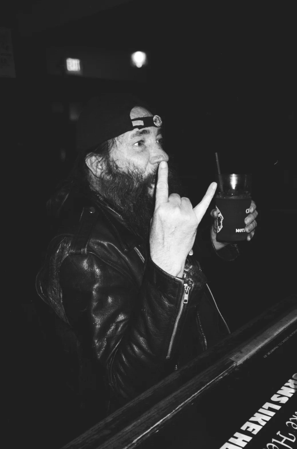 A man with a beard wearing a backward baseball cap and leather jacket, holding a drink, making a sign with his hand, in a dark setting.