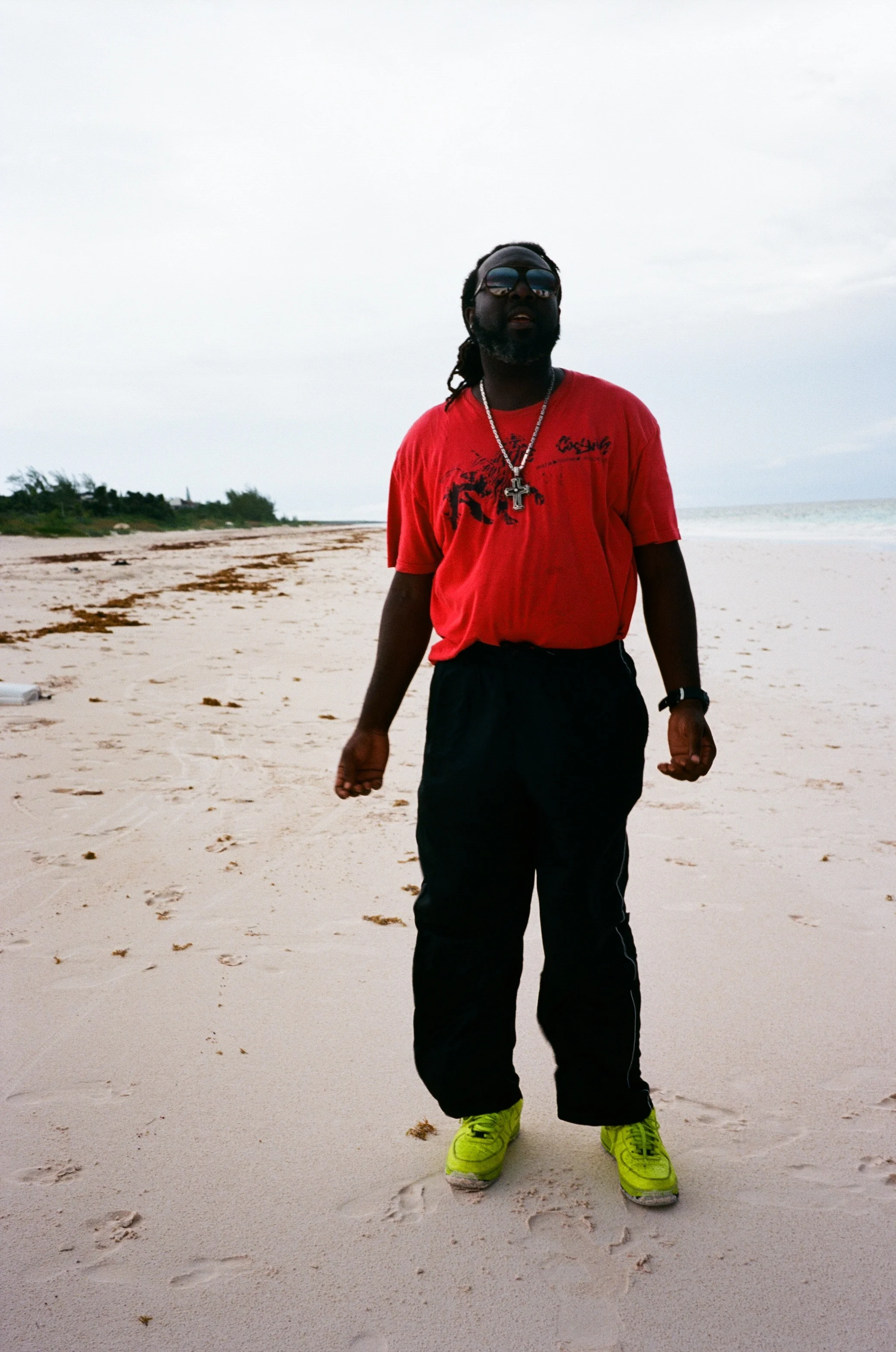 A man wearing sunglasses, a red t-shirt, black pants, and bright yellow shoes standing on a sandy beach.
