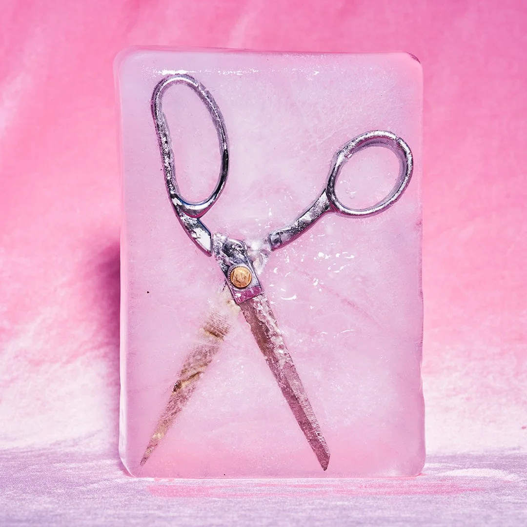 A pair of small silver scissors embedded in clear, pink resin with a gold screw in the center, set against a pink background.