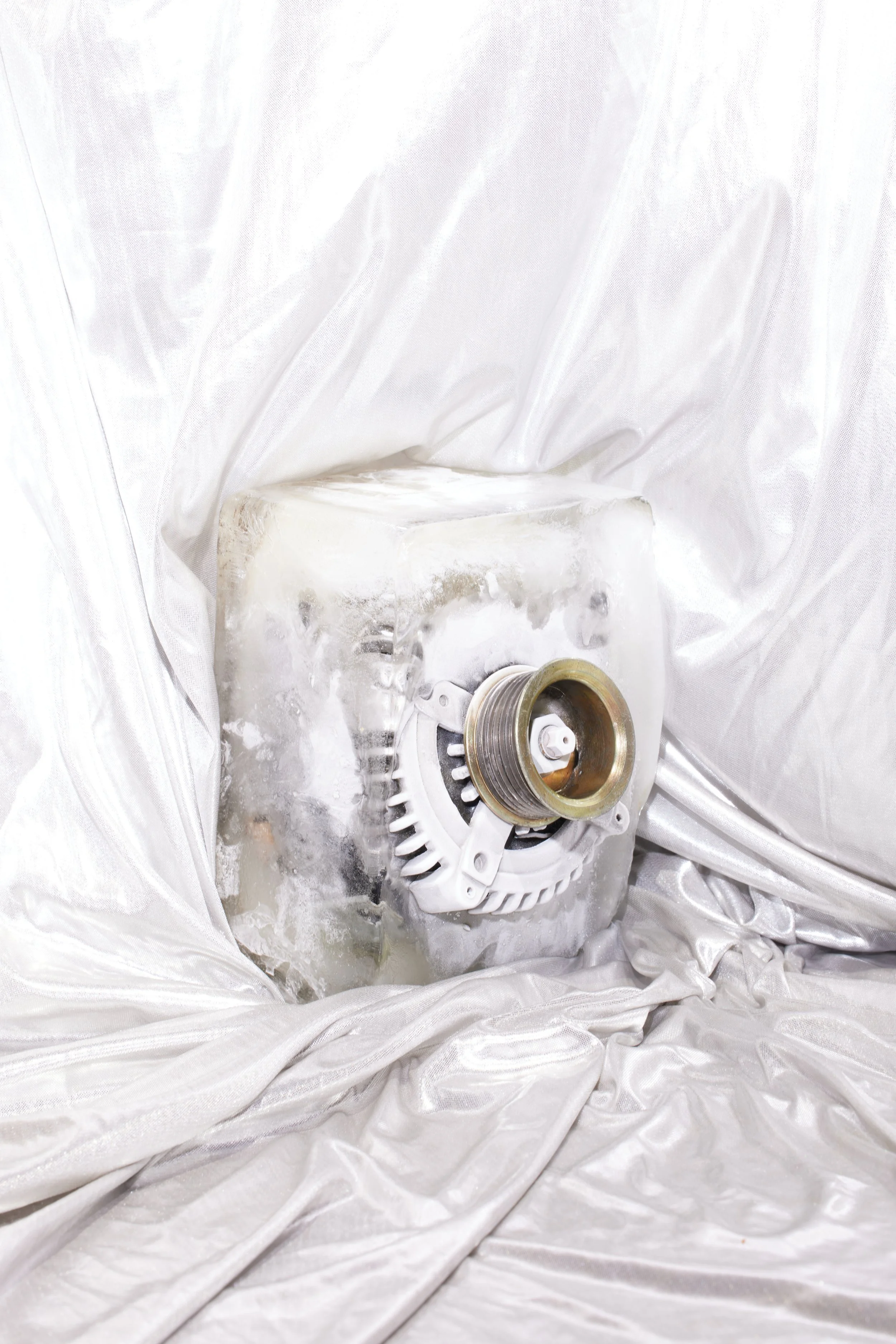 An ice sculpture resembling a machine part, with a gold-colored pulley and white fan blades, displayed on a white fabric background.