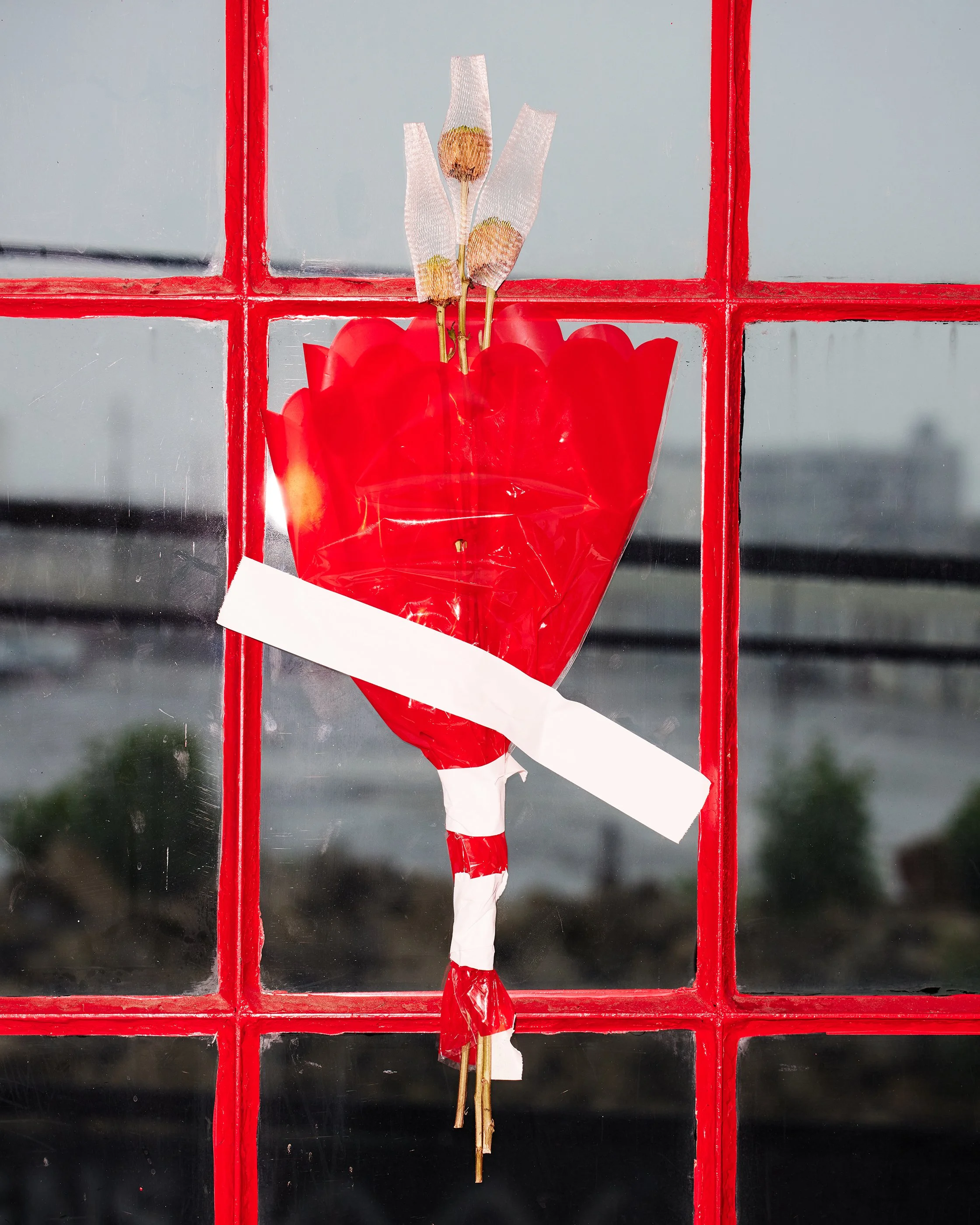 A decorative faux flower bouquet attached to a glass window with red square frames. The bouquet consists of red petals, dried seed heads, and white tape.