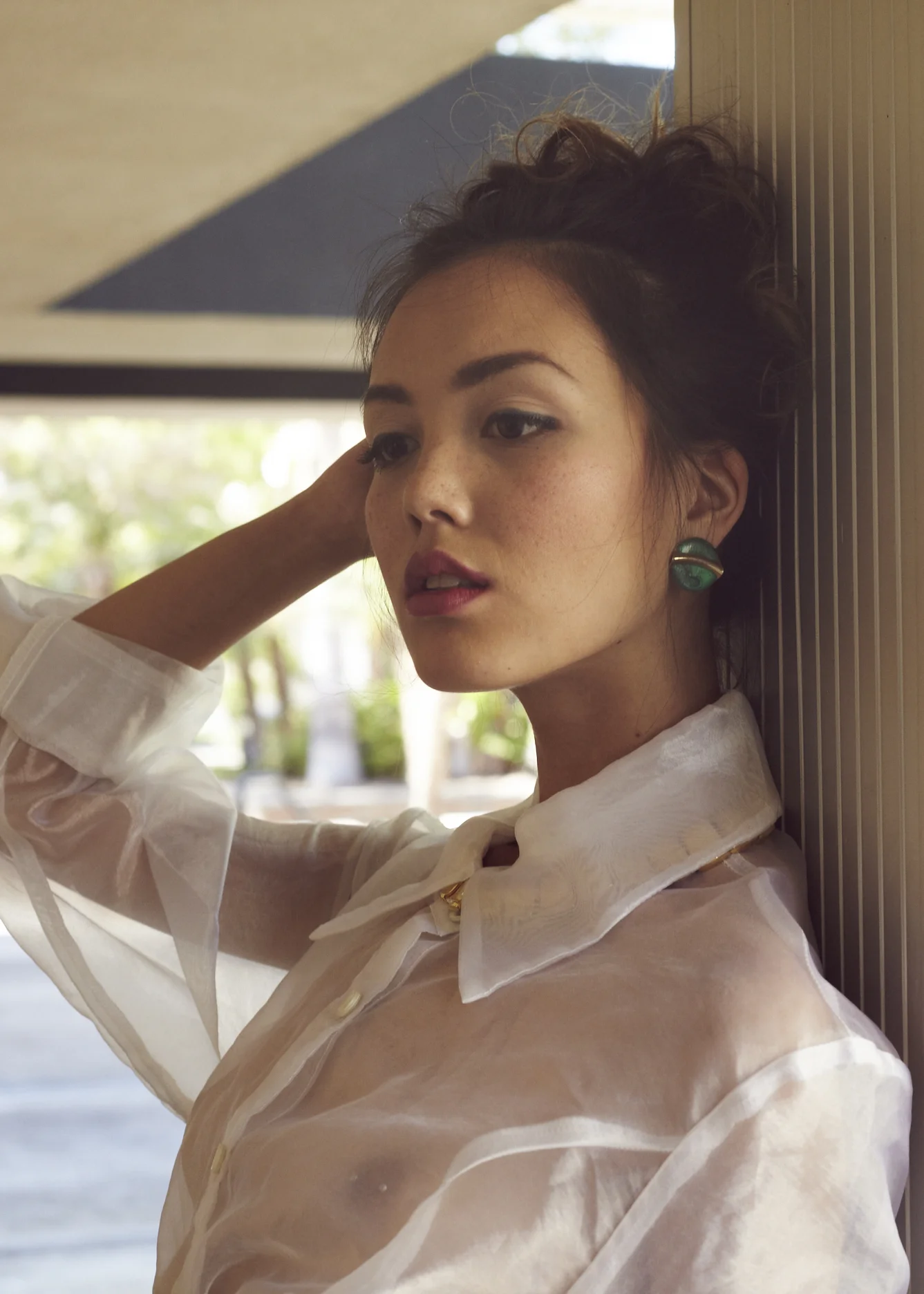A woman with dark hair and large green earrings, dressed in a sheer white blouse, leaning against a wall, with a thoughtful expression, indoors with natural light.