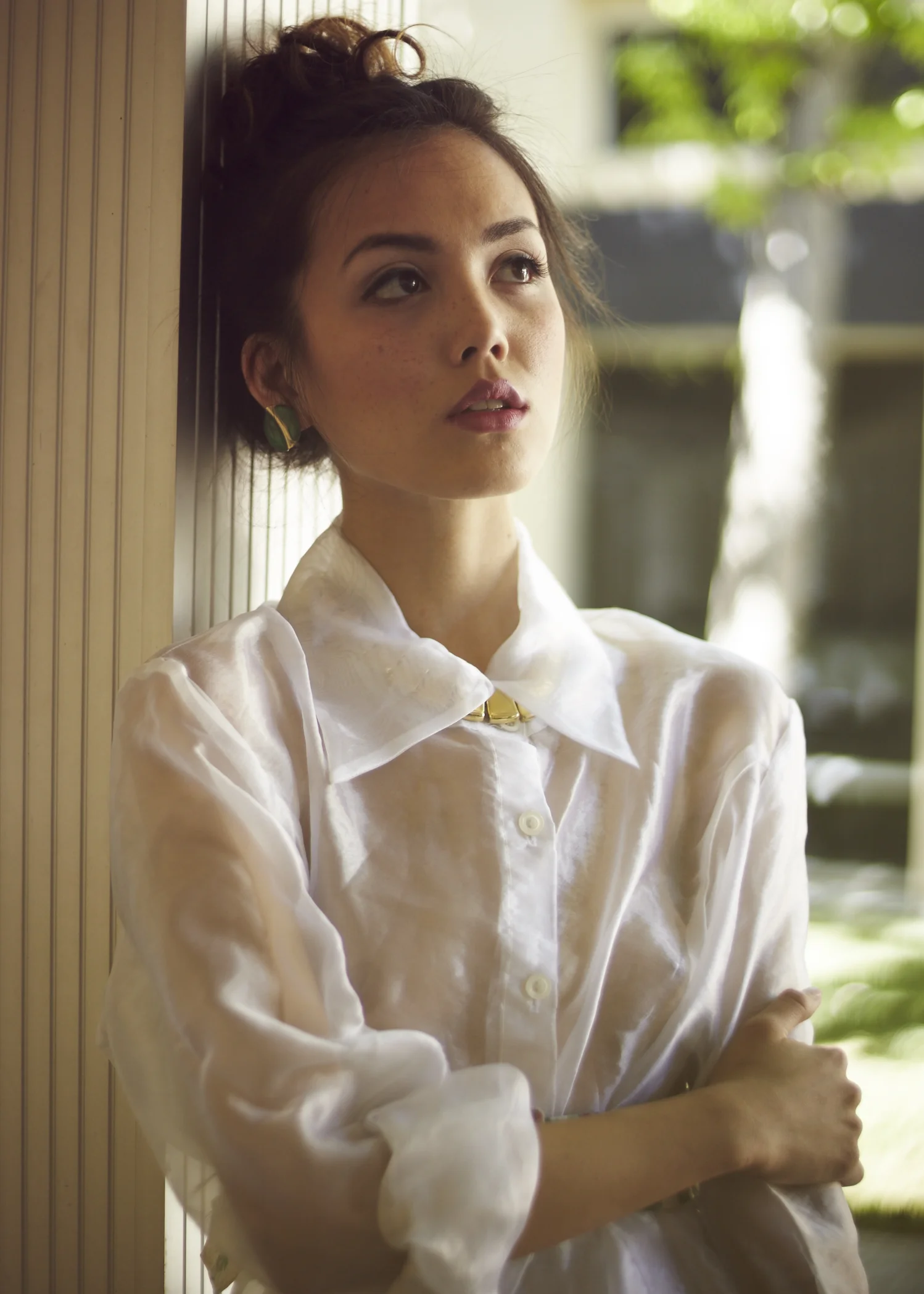 A woman with dark hair styled in a messy updo, wearing a white satin blouse, standing with arms crossed against a wooden wall, looking thoughtfully to her right, with blurred greenery outside the window in the background.