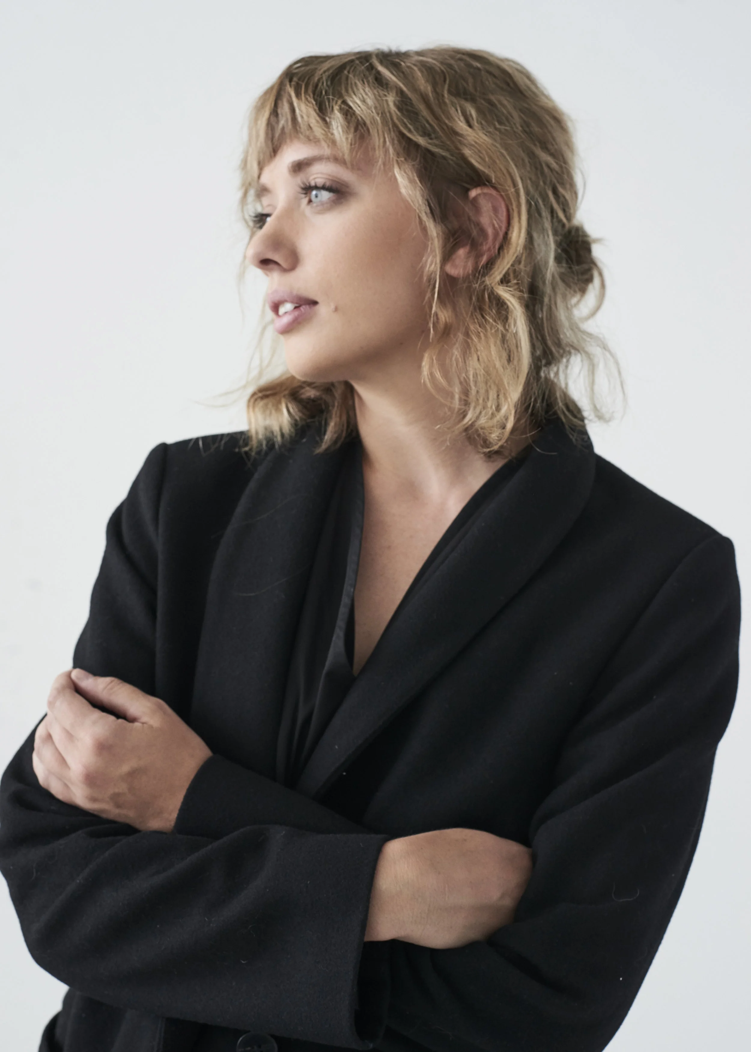 A woman with blonde, wavy hair wearing a black blazer, looking to her left with crossed arms.