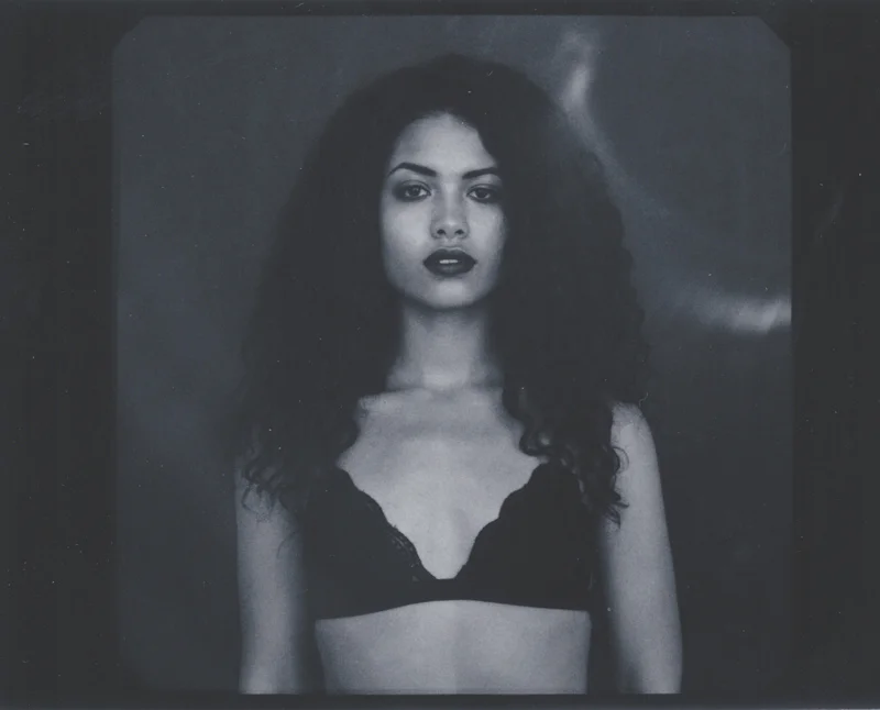 Black and white photo of a woman with curly hair, wearing a black lace bra, looking directly at the camera with a serious expression.