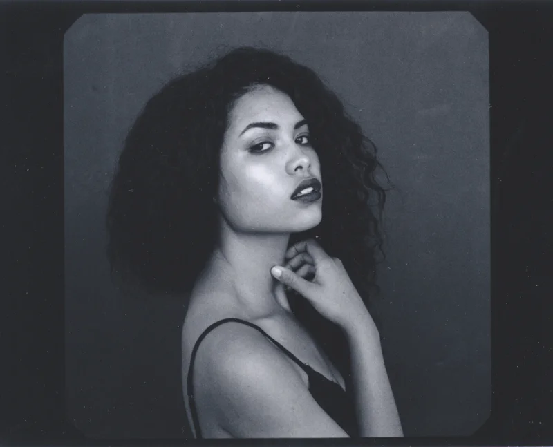 Black and white portrait of a woman with dark, curly hair, wearing a spaghetti strap top, looking over her shoulder with a confident expression, against a plain background.