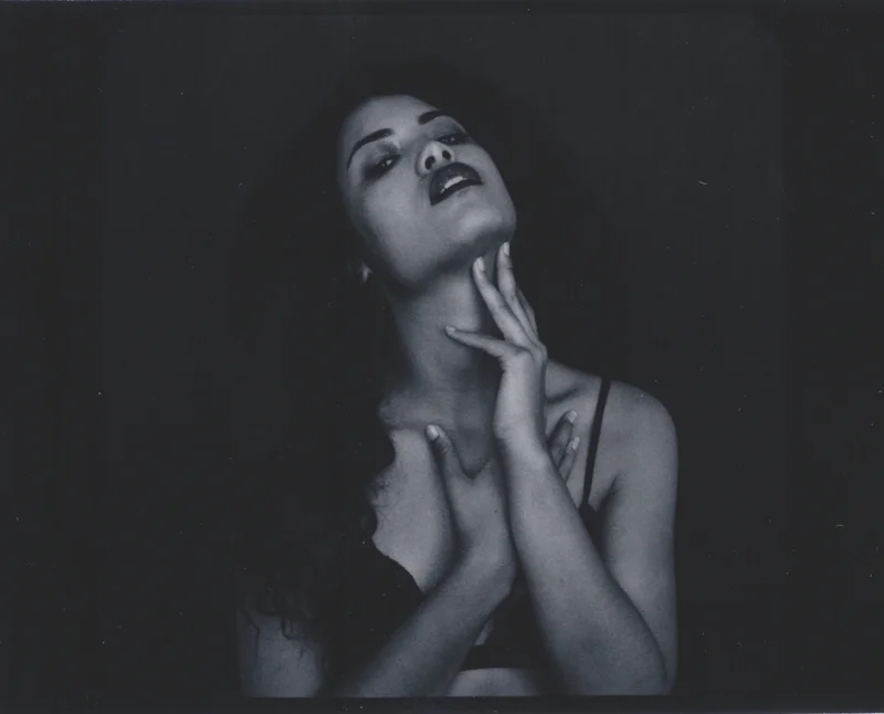 A black and white portrait of a woman with dark, curly hair, wearing dark lipstick. She is looking slightly upwards with her eyes half-closed, one hand resting on her neck and the other touching her chest, with a sensual expression.