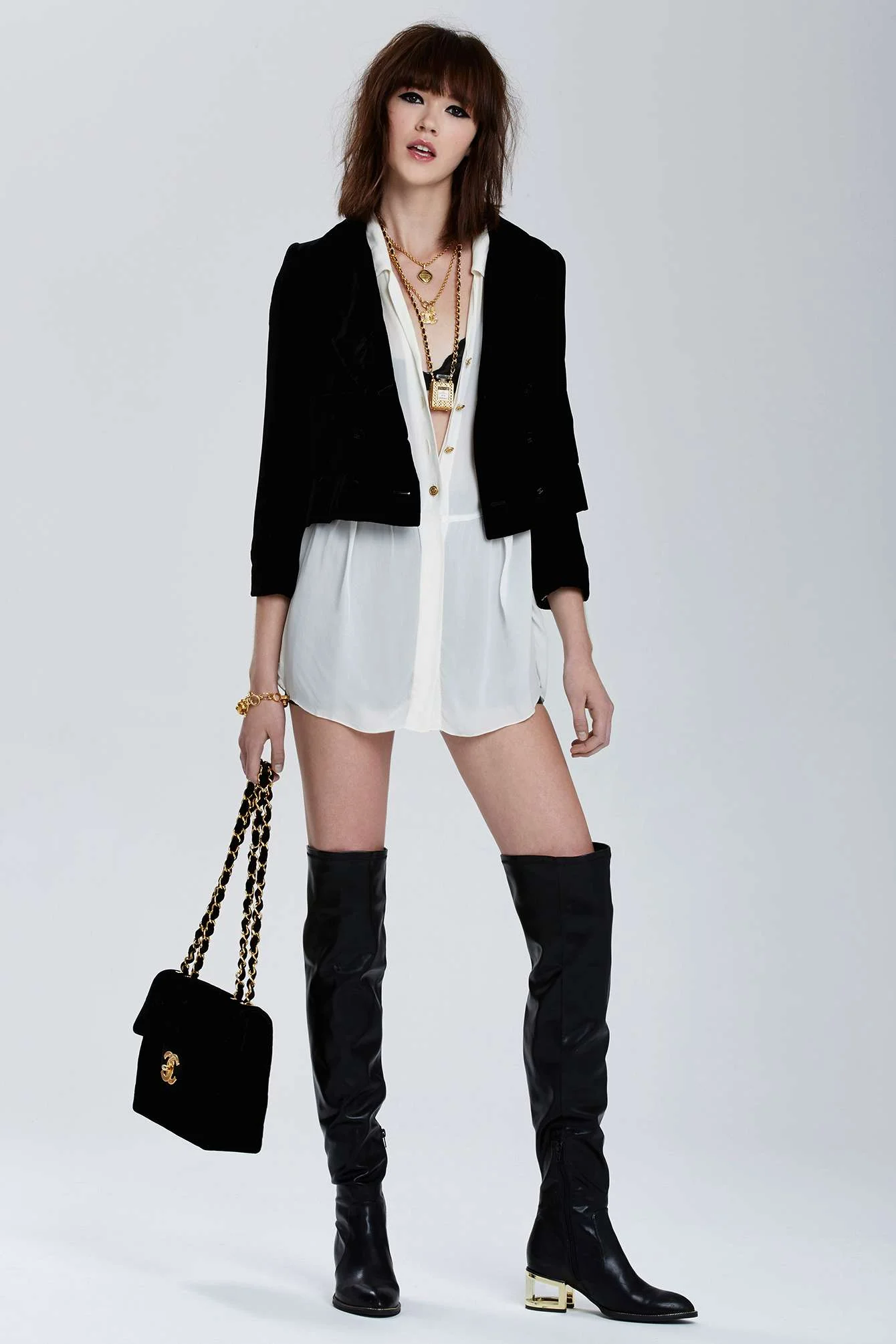 Fashion model wearing a white dress, black jacket, black thigh-high boots with heels, holding a black purse, and accessorized with gold jewelry.