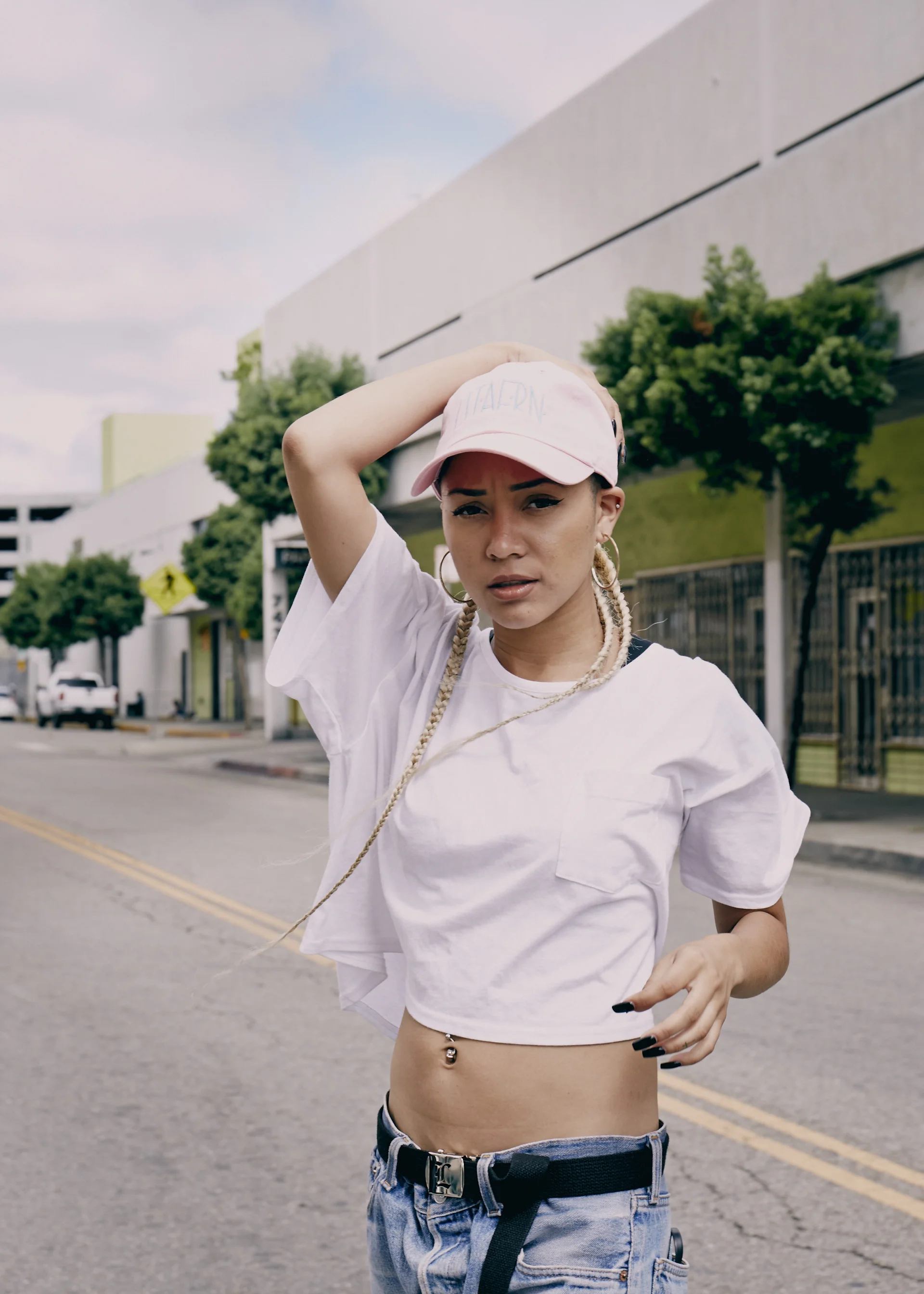 A young woman standing on the street, wearing a pink cap, white cropped T-shirt, and ripped jeans, with one hand on her head and looking at the camera.