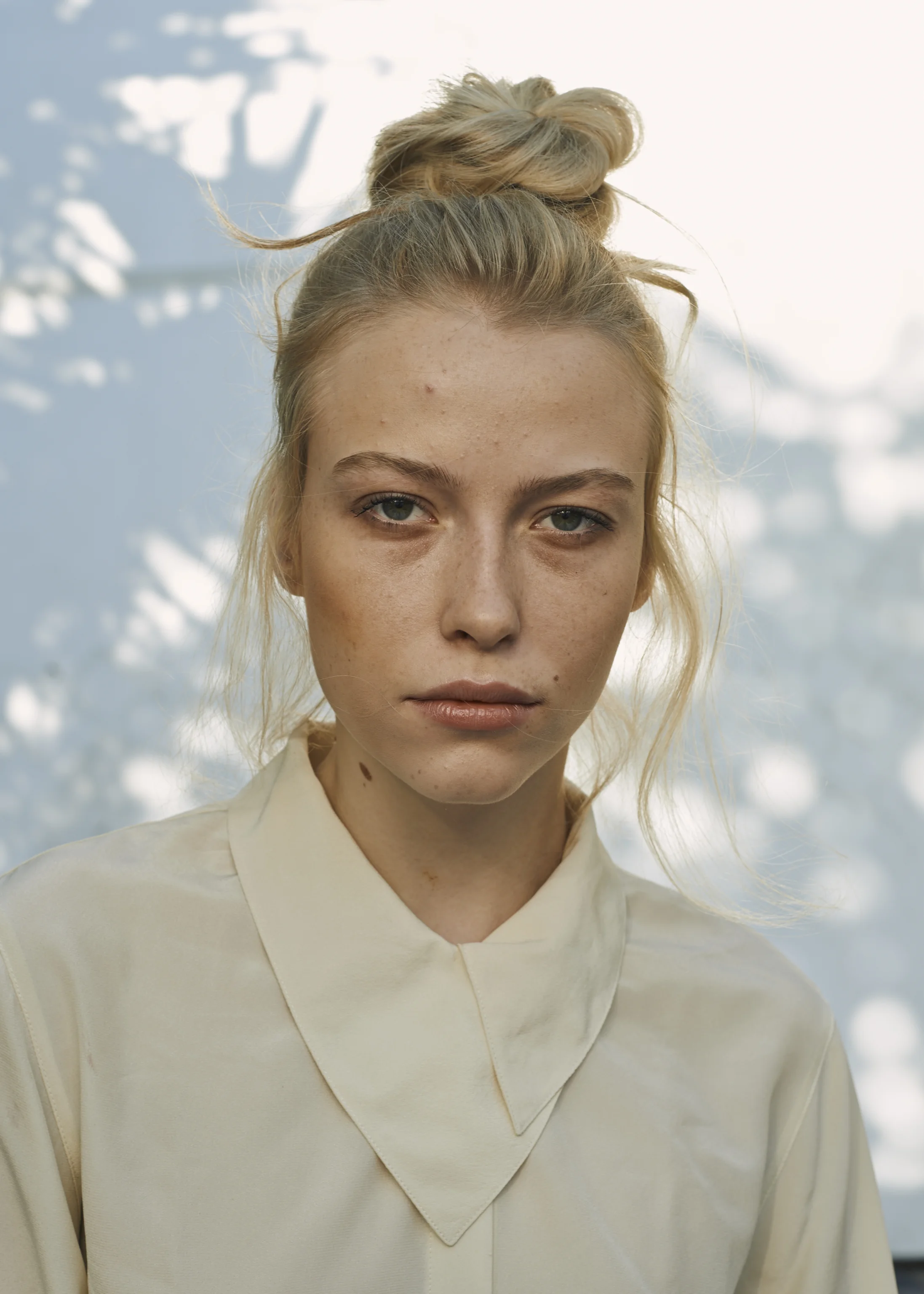 Close-up of a young woman with blonde hair in a messy bun, wearing a cream-colored blouse, standing outdoors with blurred trees in the background.