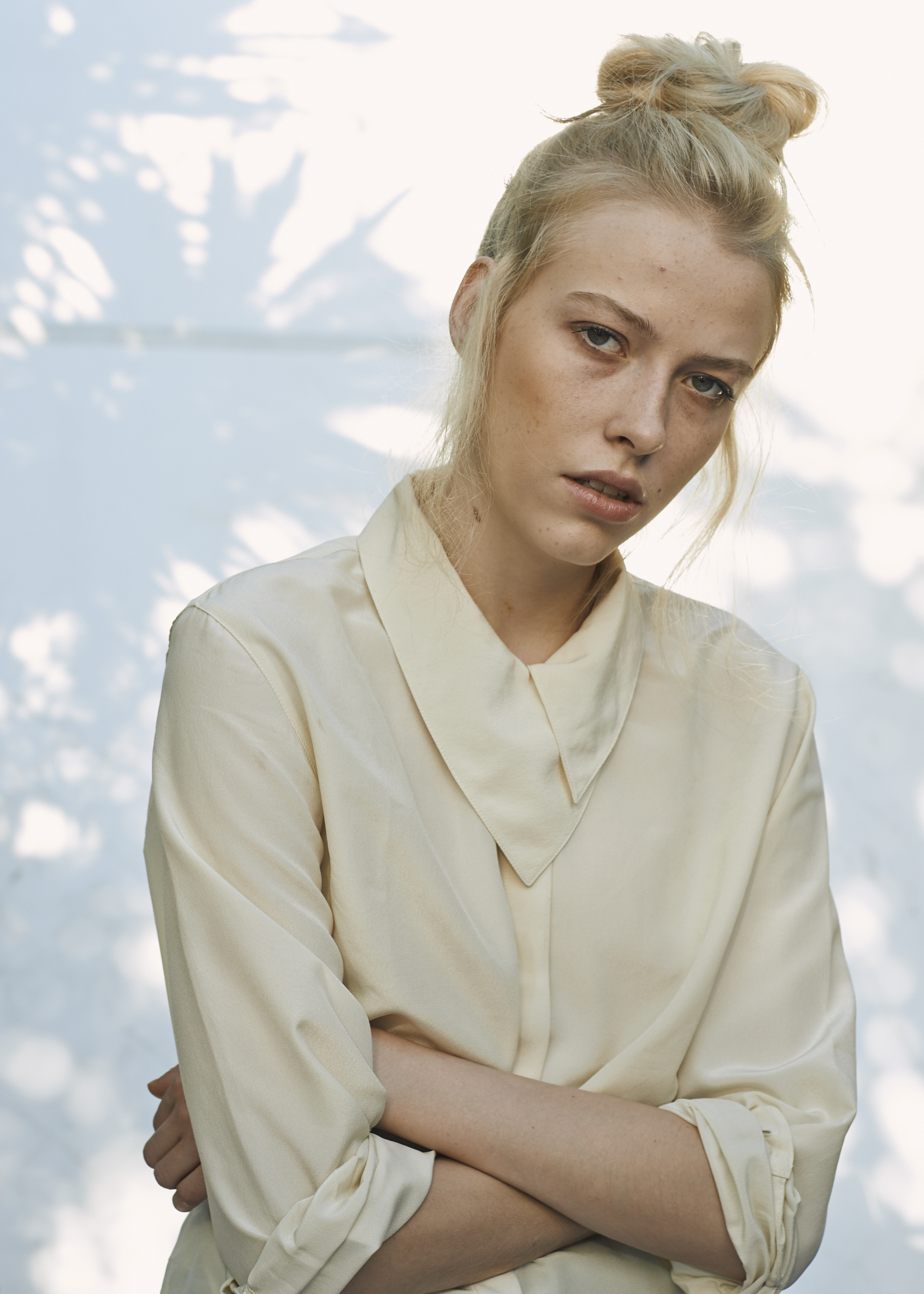 A young woman with blond hair in a messy bun, wearing a light-colored satin blouse, crosses her arms, with a background of blurred tree shadows and sky.