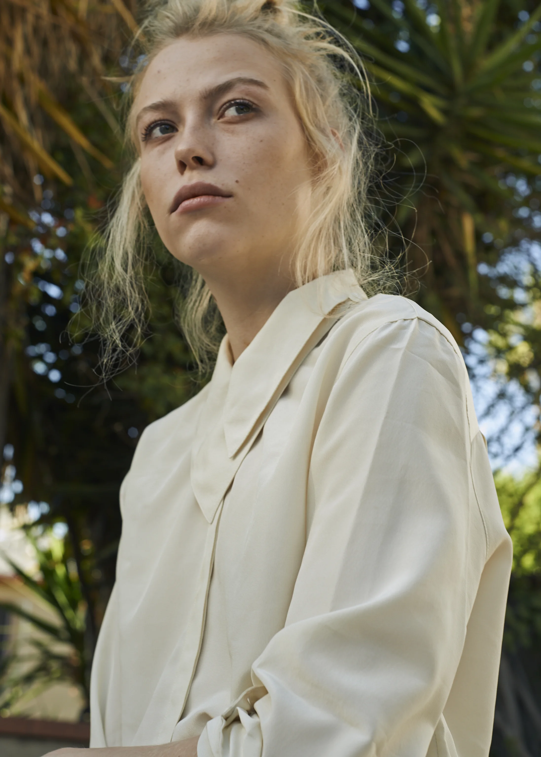 A young woman with blonde hair and blue eyes looking slightly away, wearing a cream-colored blouse, outdoors with green trees and blue sky in background.
