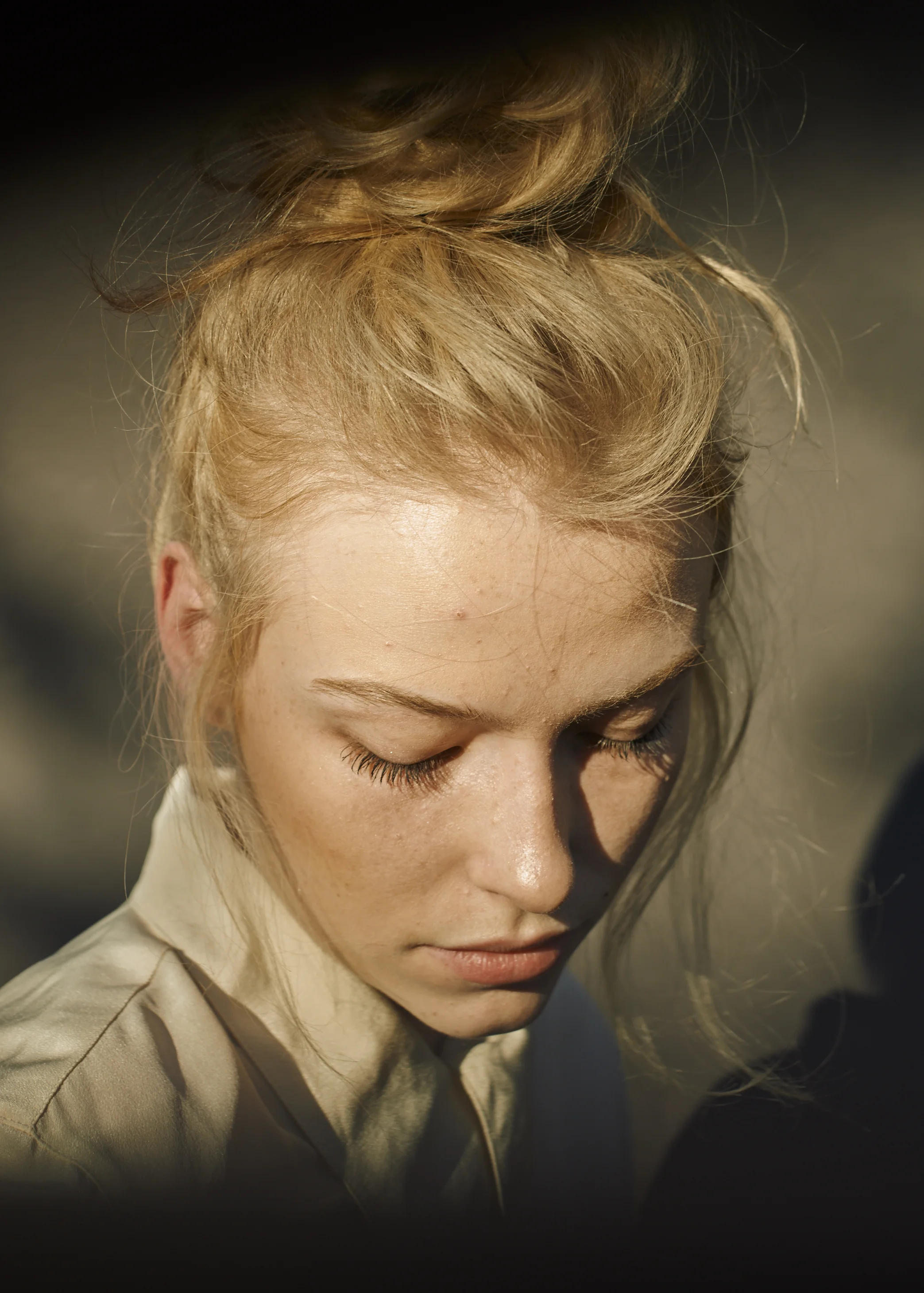 Close-up of a young woman with red hair styled in an updo, looking downward with her eyes closed, lit by warm sunlight.
