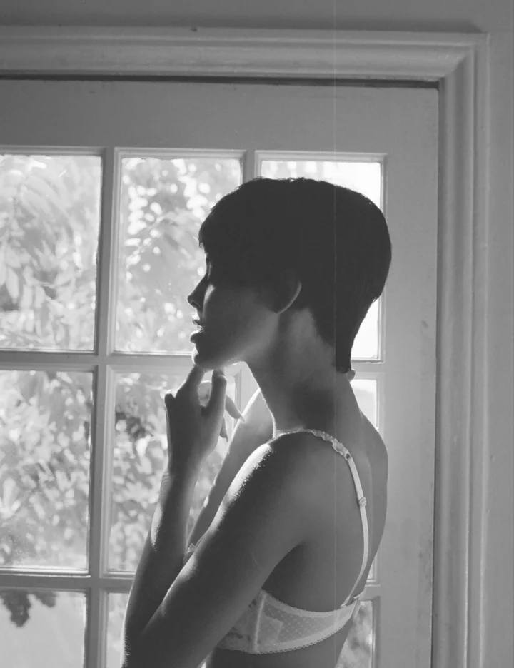 A silhouette of a woman with short hair standing in profile near a window, touching her chin with her finger, dressed in a tank top, backlit by natural light from outside.
