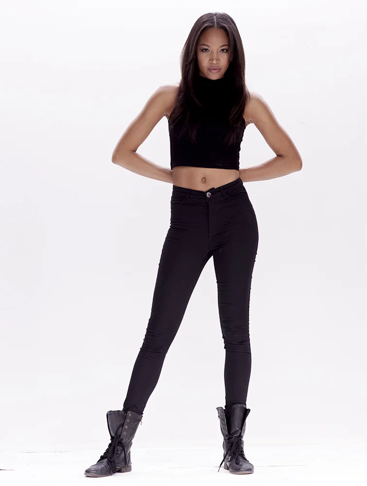 A young woman with long dark hair wearing a sleeveless black crop top, high-waisted black pants, and black lace-up boots standing with hands on hips against a plain white background.