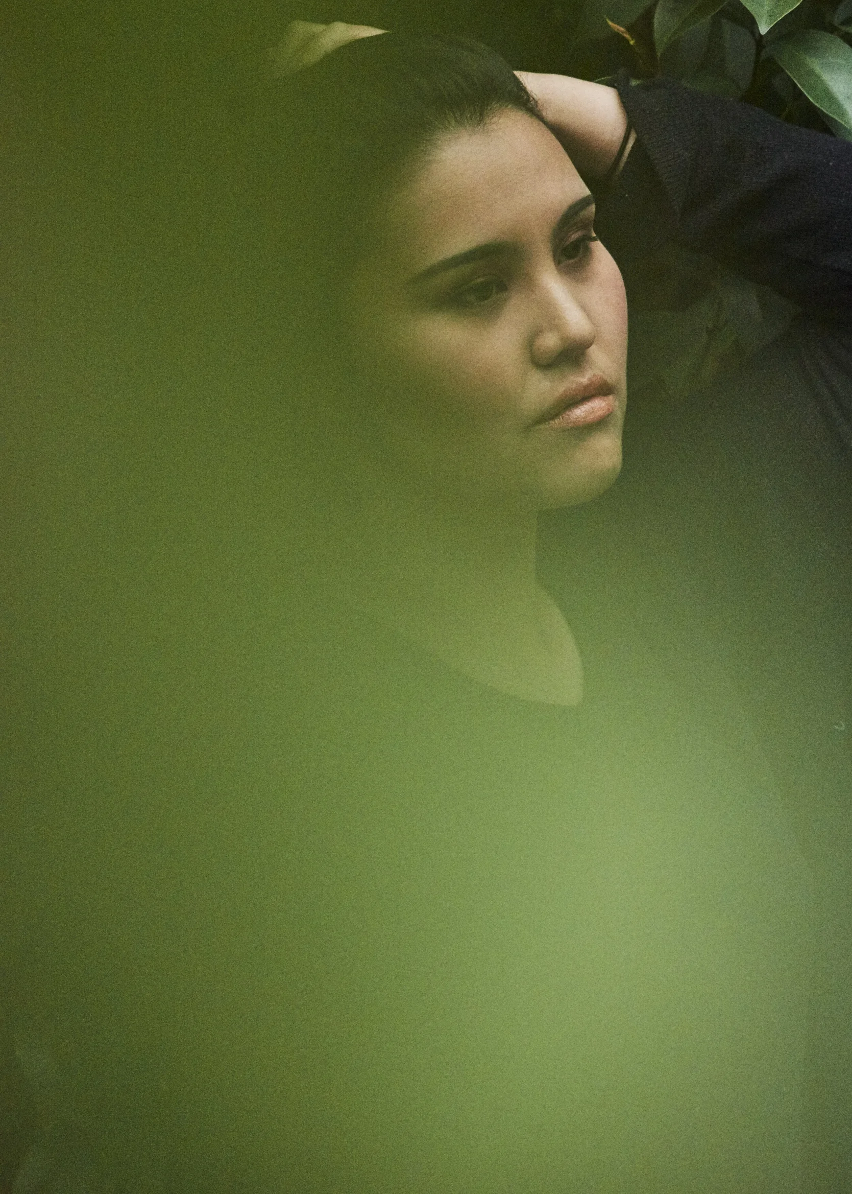 A woman with dark hair and makeup, posing with her hand behind her head, partially obscured by greenish blur in front of the camera.