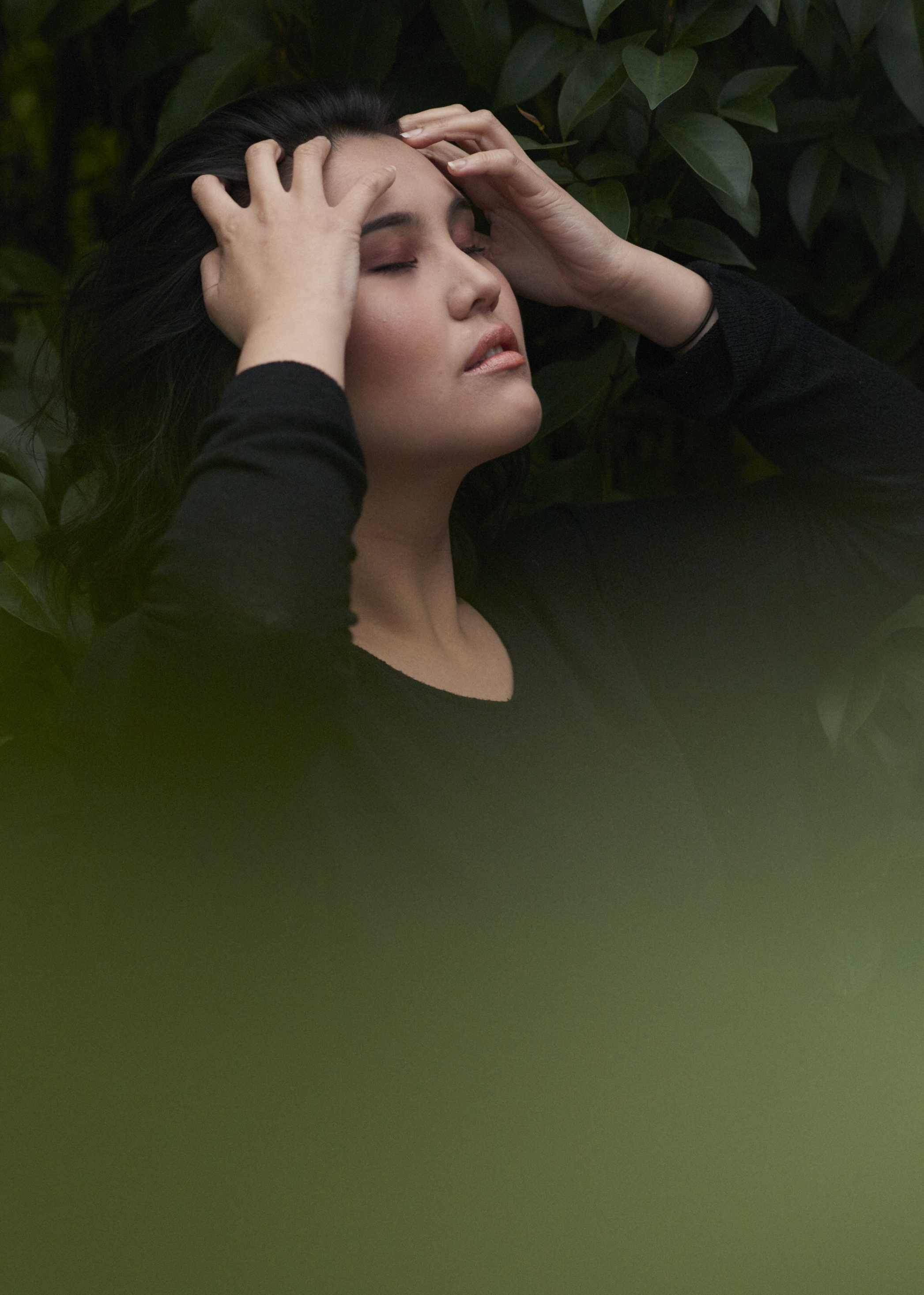 A woman with dark hair and closed eyes, holding her head with both hands, surrounded by green leaves.
