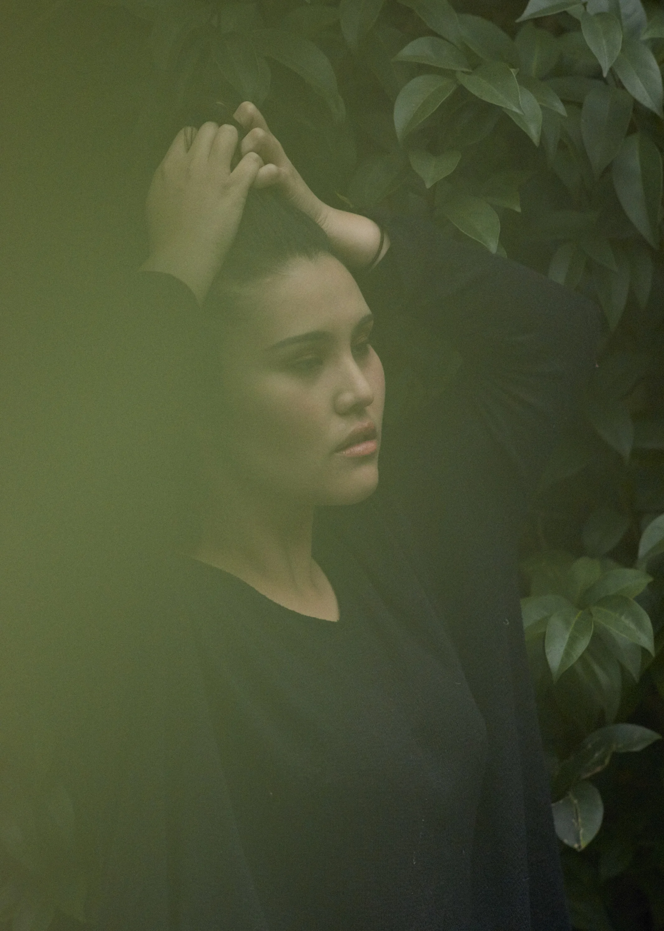 A woman with dark hair and makeup, wearing a black top, lies among green foliage, looking contemplatively to the side.