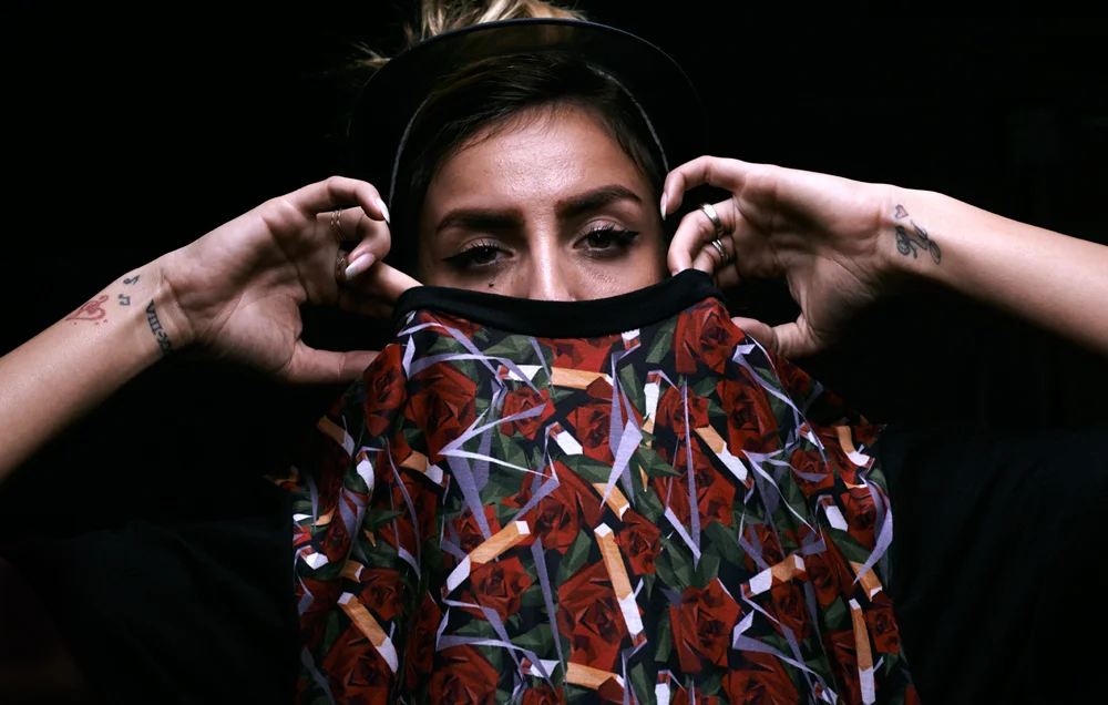 A woman with tattoos on her arms is adjusting a floral face mask with red roses and green leaves, covering her nose and mouth, against a dark background.