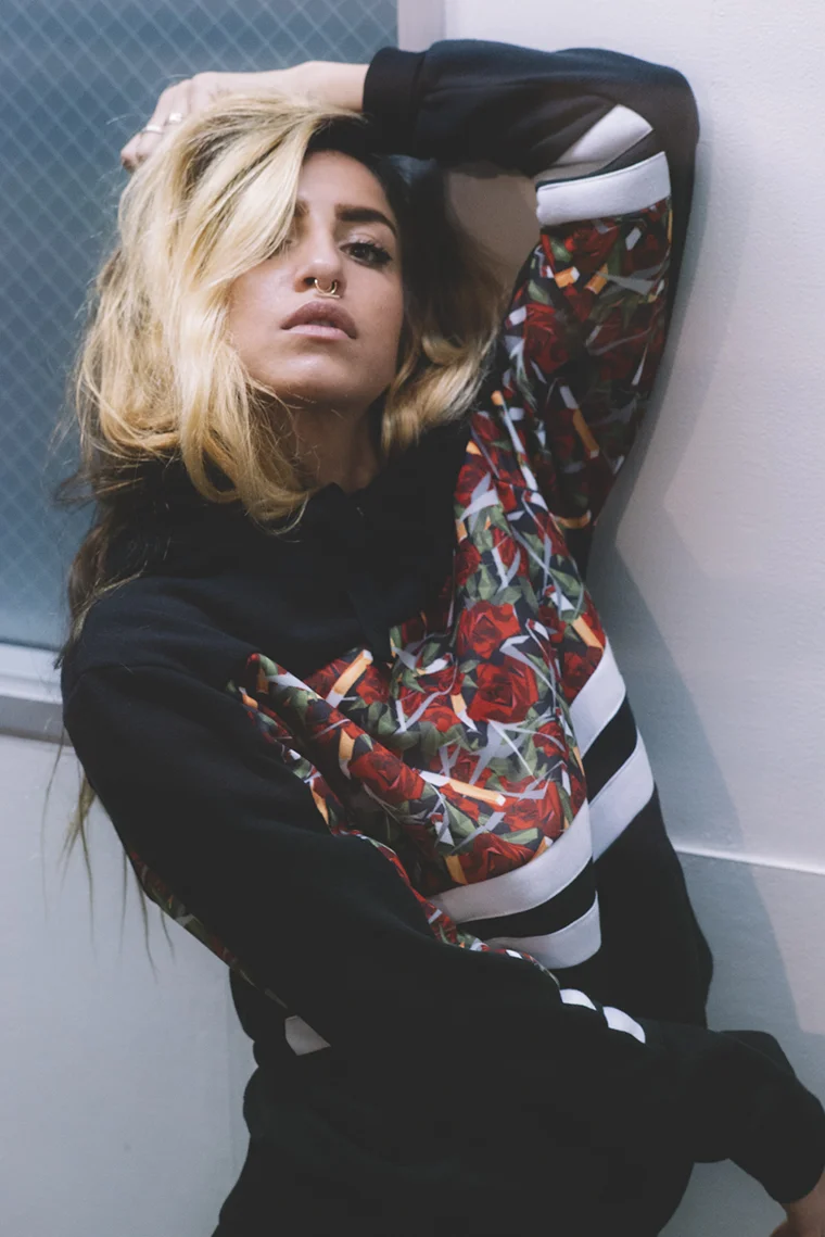 A woman with blonde, wavy hair and a septum nose piercing is posing with her arm over her head, leaning against a wall inside a building. She is wearing a black hoodie with a colorful floral pattern on the sleeves and a white stripe detail. The backg