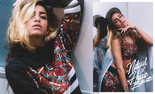 A woman with blonde and dark hair, wearing a colorful patterned dress, posing stylishly with her hand near her face, and her reflection in a mirror featuring a white graphic text that reads "Natural & Funz BELZ."