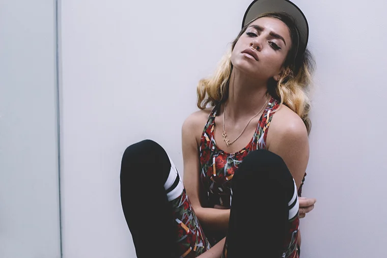 Young woman sitting on the floor with arms crossed, wearing a colorful sleeveless top, black pants with white stripes, a black cap, and gold jewelry, against a plain white wall.