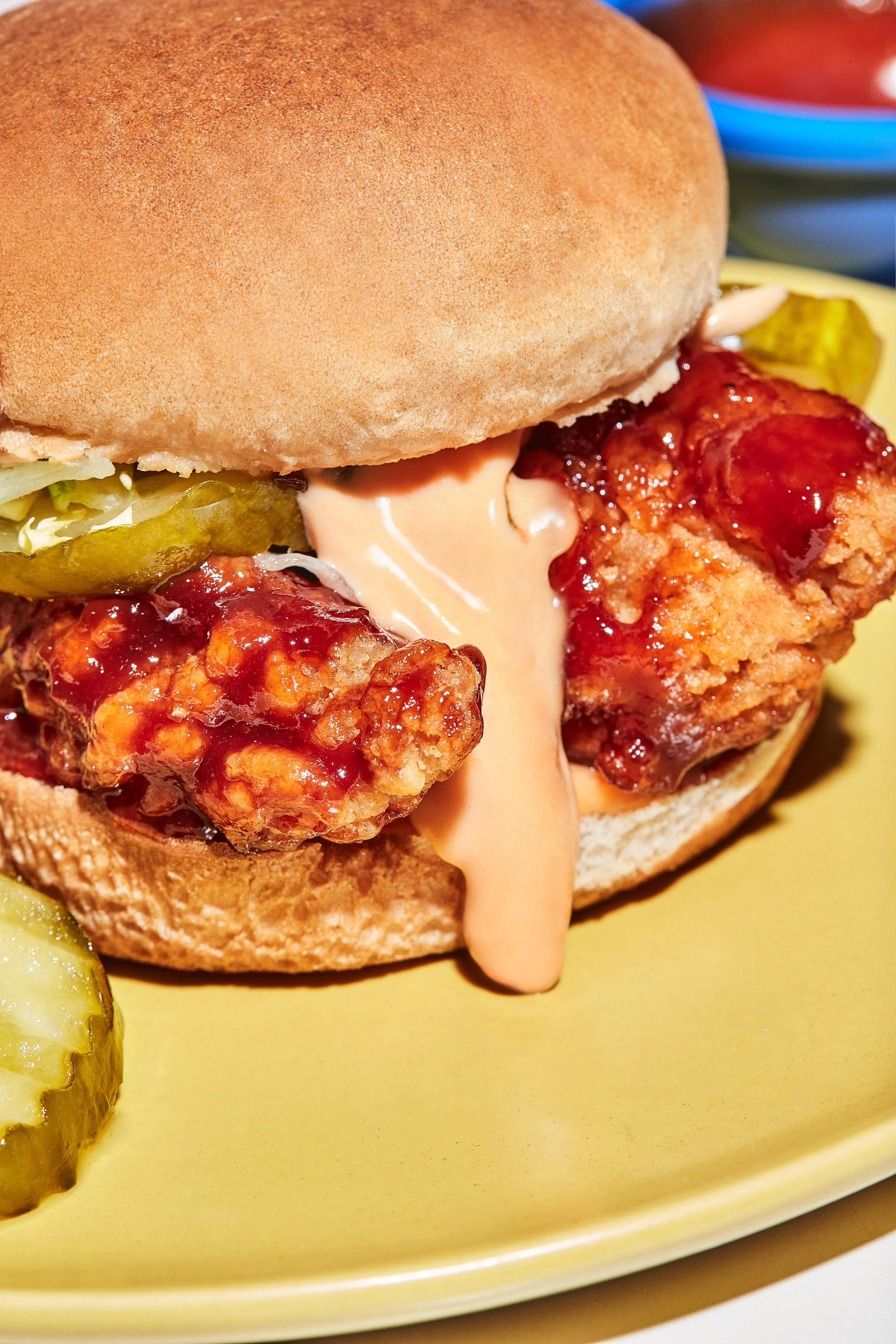 Close-up of fried chicken sandwich with mayonnaise, pickles, and hot sauce on a yellow plate.