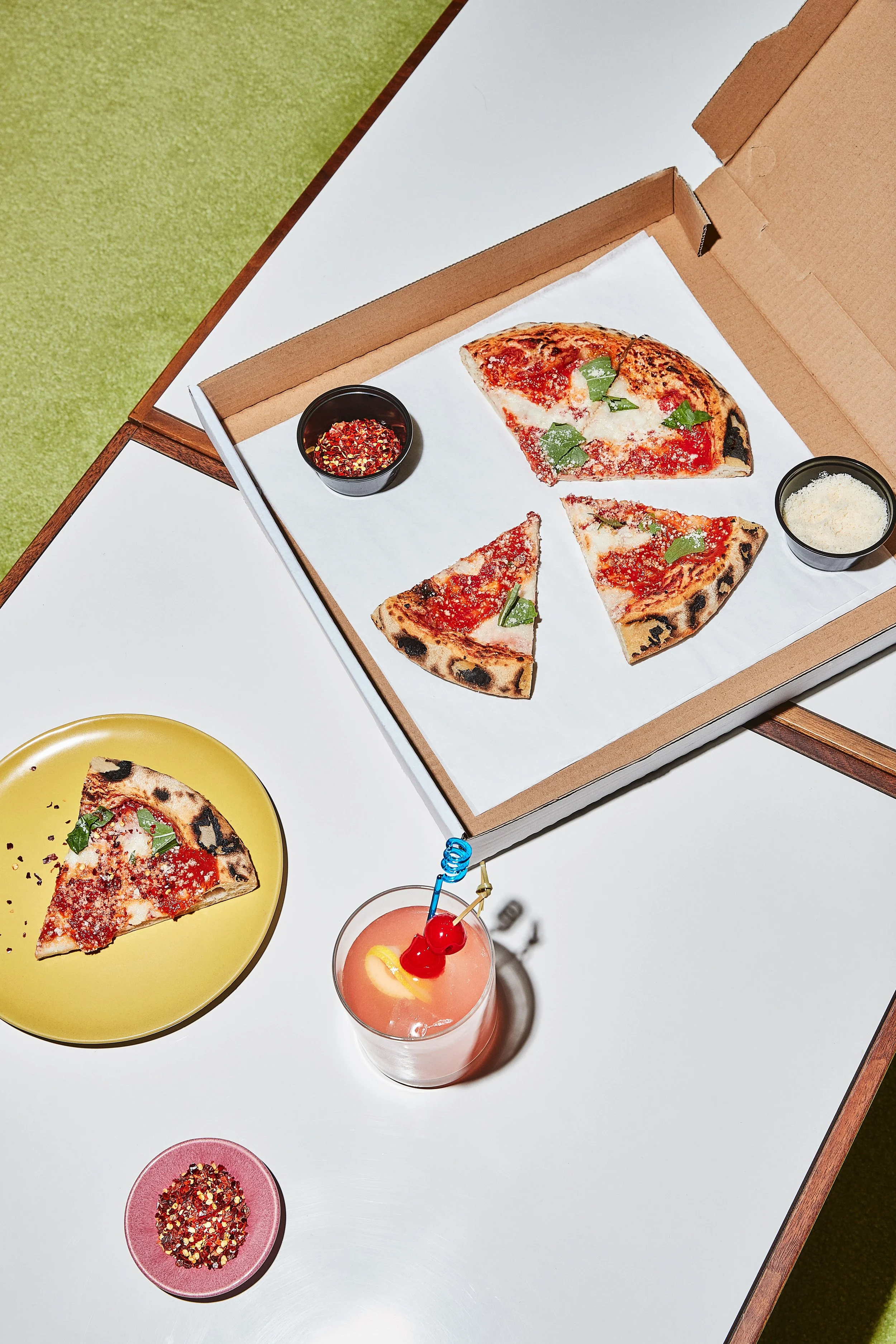 A box of three pizza slices with basil leaves, a yellow plate with one pizza slice, a pink bowl of colorful seasoning, and a pink cocktail with cherries and lemon on a white table.