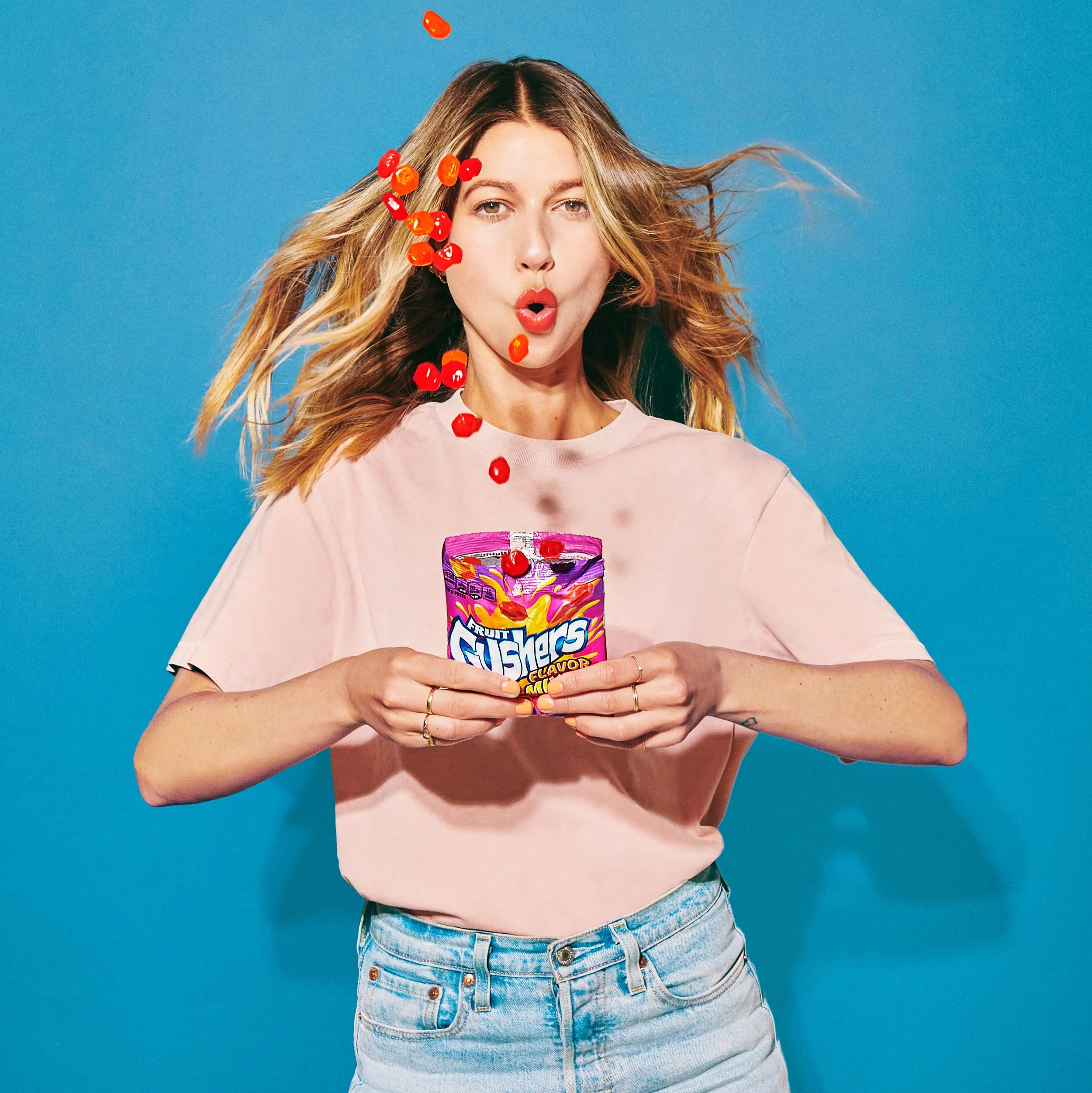 Young woman with blonde hair wearing a pink T-shirt and blue jeans, holding a bag of Fruit Gushers candy, with cherry pieces falling around her in front of a blue background.